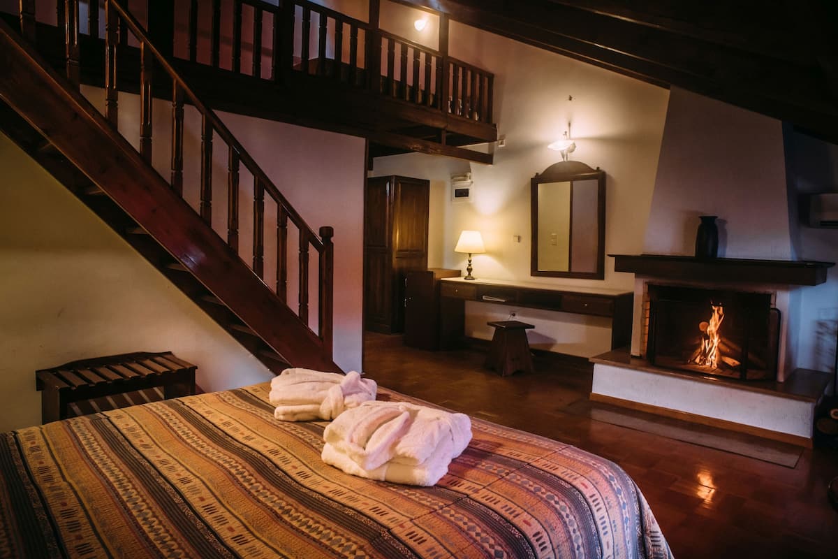 A hotel room with a wooden staircase, a fireplace, a bed with patterned bedding, and a nightstand with a lamp.