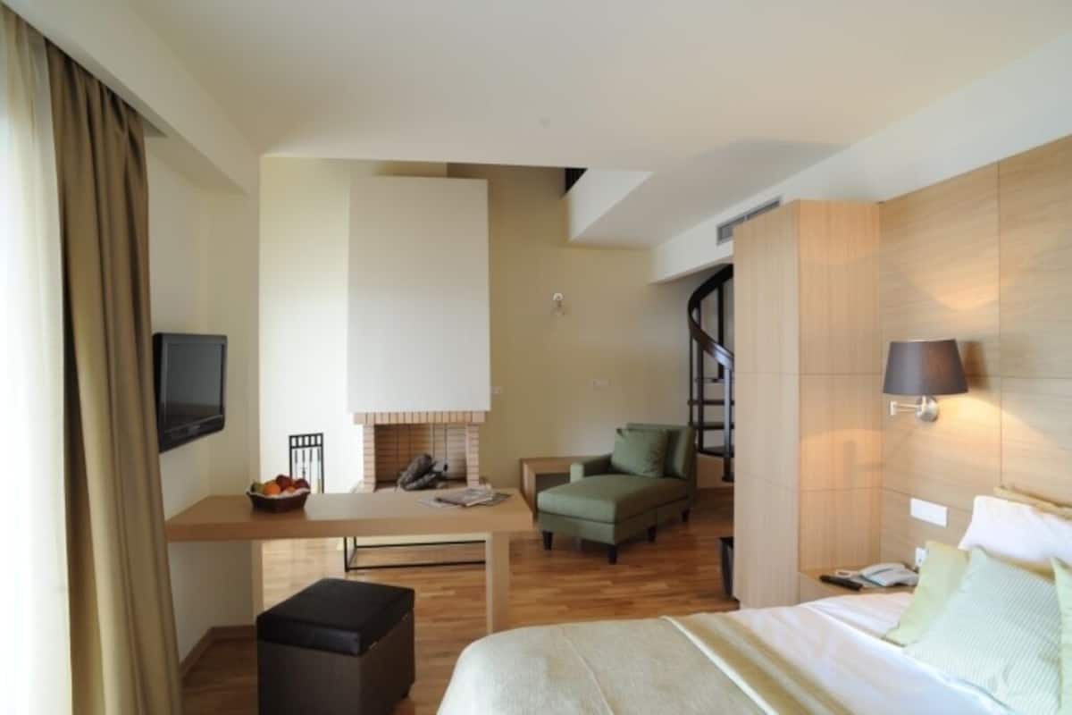 A hotel room with a bed, a desk, a chair, a sofa, a TV, a lamp, and a fireplace.