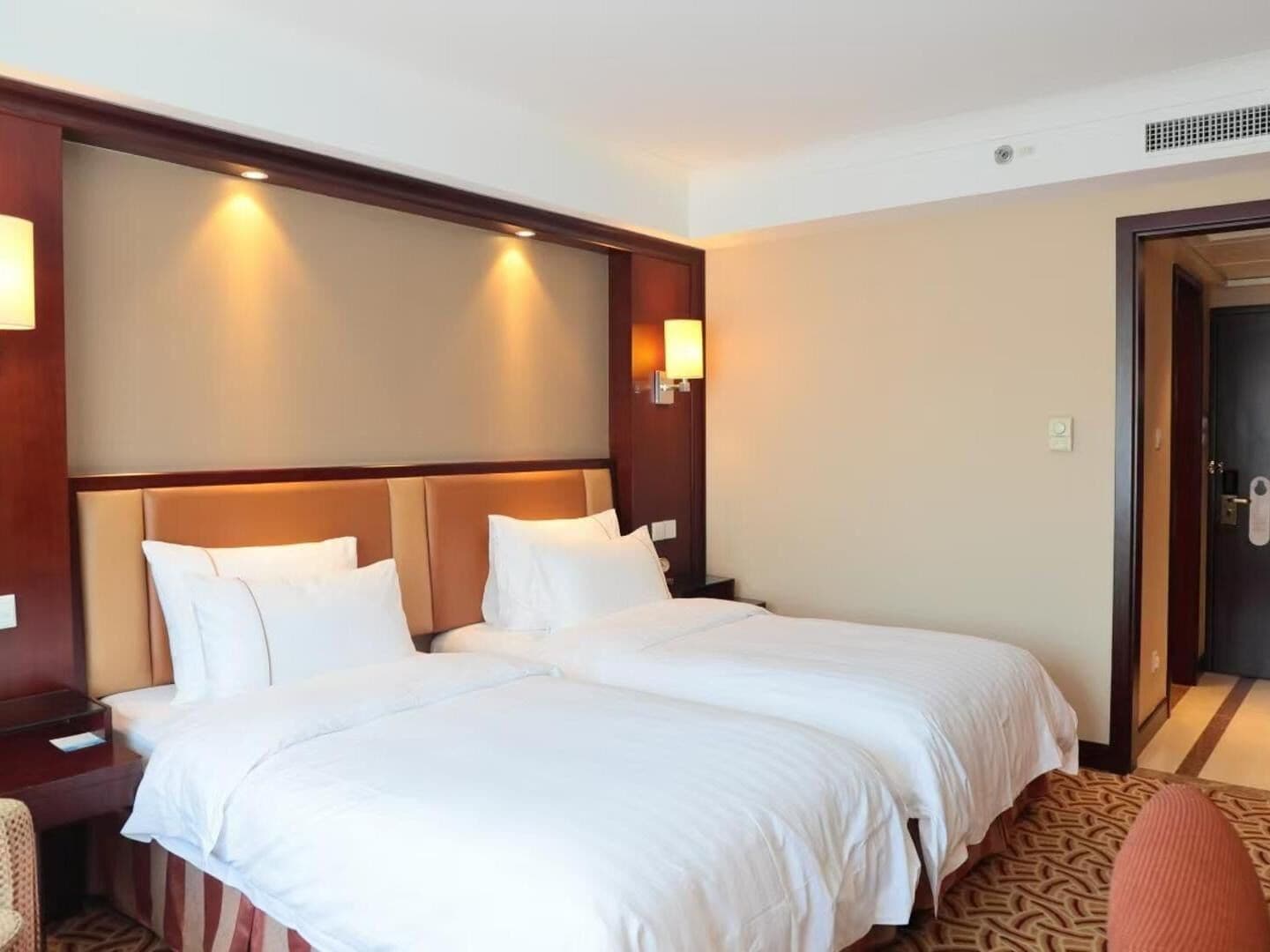 Een hotelkamer met twee bedden, een houten hoofdbord, nachtlampjes, een kamerbreed tapijt met een patroon, en een deur aan de rechterkant.