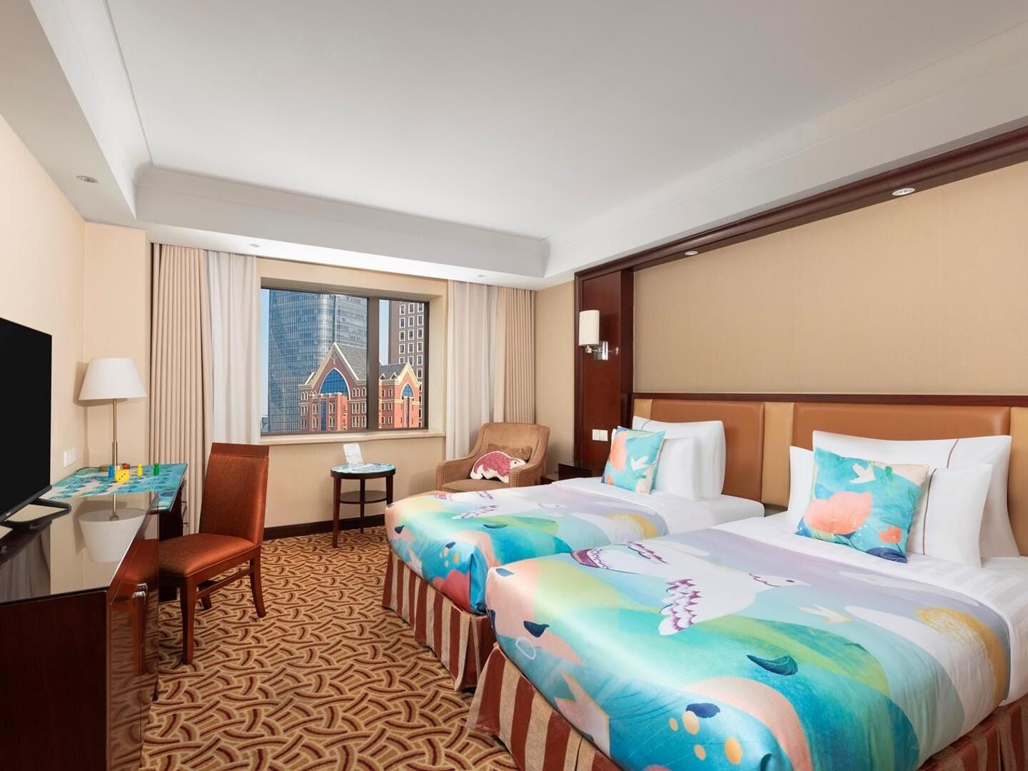 A hotel room with two beds, a desk, a chair, a TV, and a window with a cityscape view.
