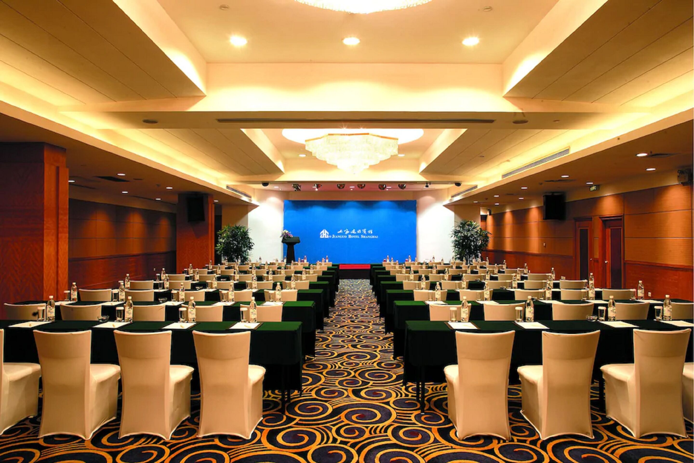 Un salón de conferencias con mesas y sillas dispuestas en filas, un fondo azul con texto y una alfombra estampada.
