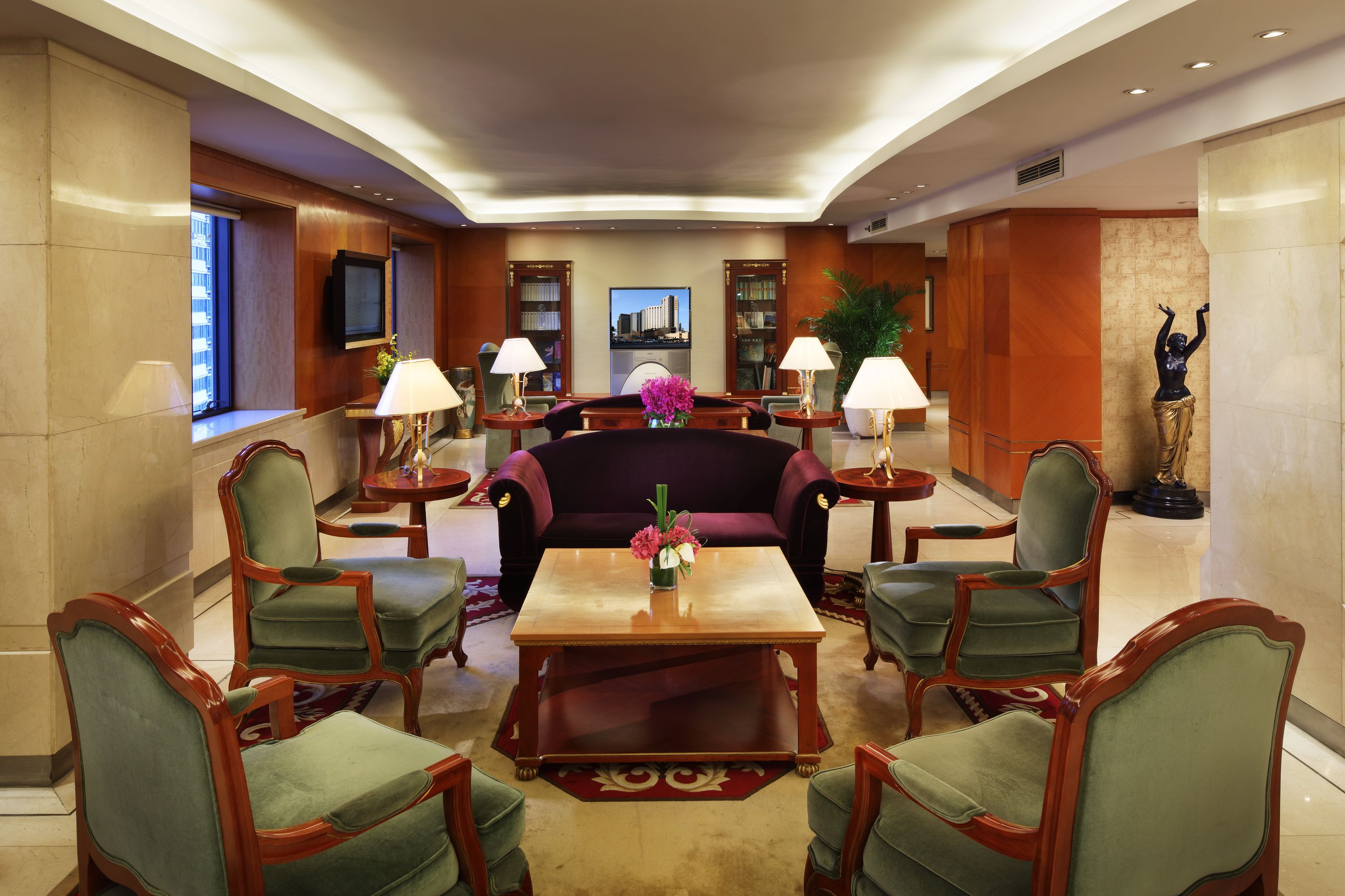 A lobby with a dark-colored sofa, green upholstered chairs, a wooden coffee table, and a television on the wall.