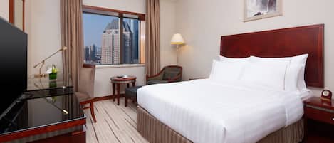 A hotel room with a large bed, a desk with a TV, a chair, a small table, and a window with a city view.