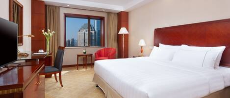 A hotel room with a large bed, a desk with a TV, a chair, a small table, and a window with a city view.