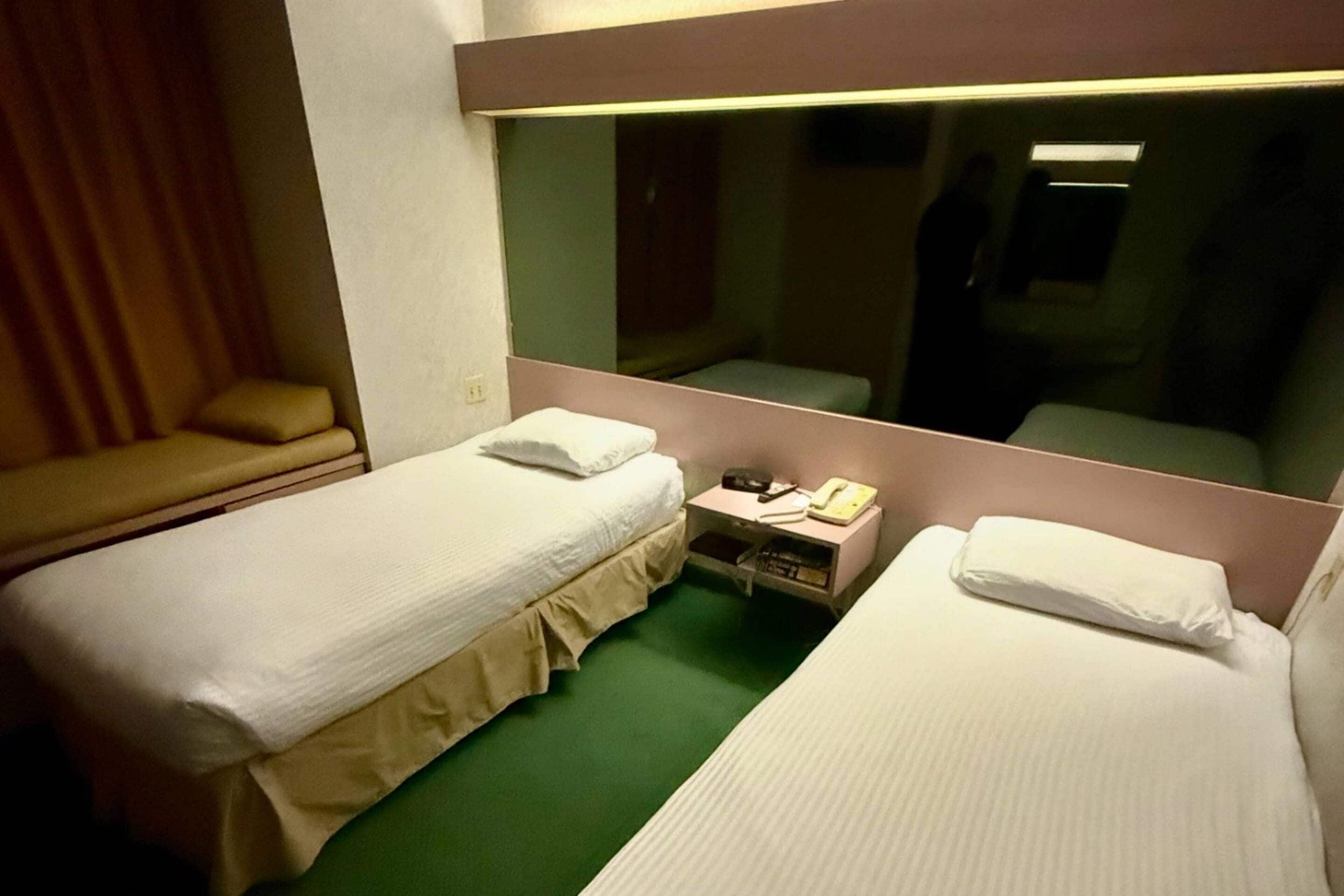 A hotel room with two single beds, a bedside table with a lamp, a mirror, and a green carpet.