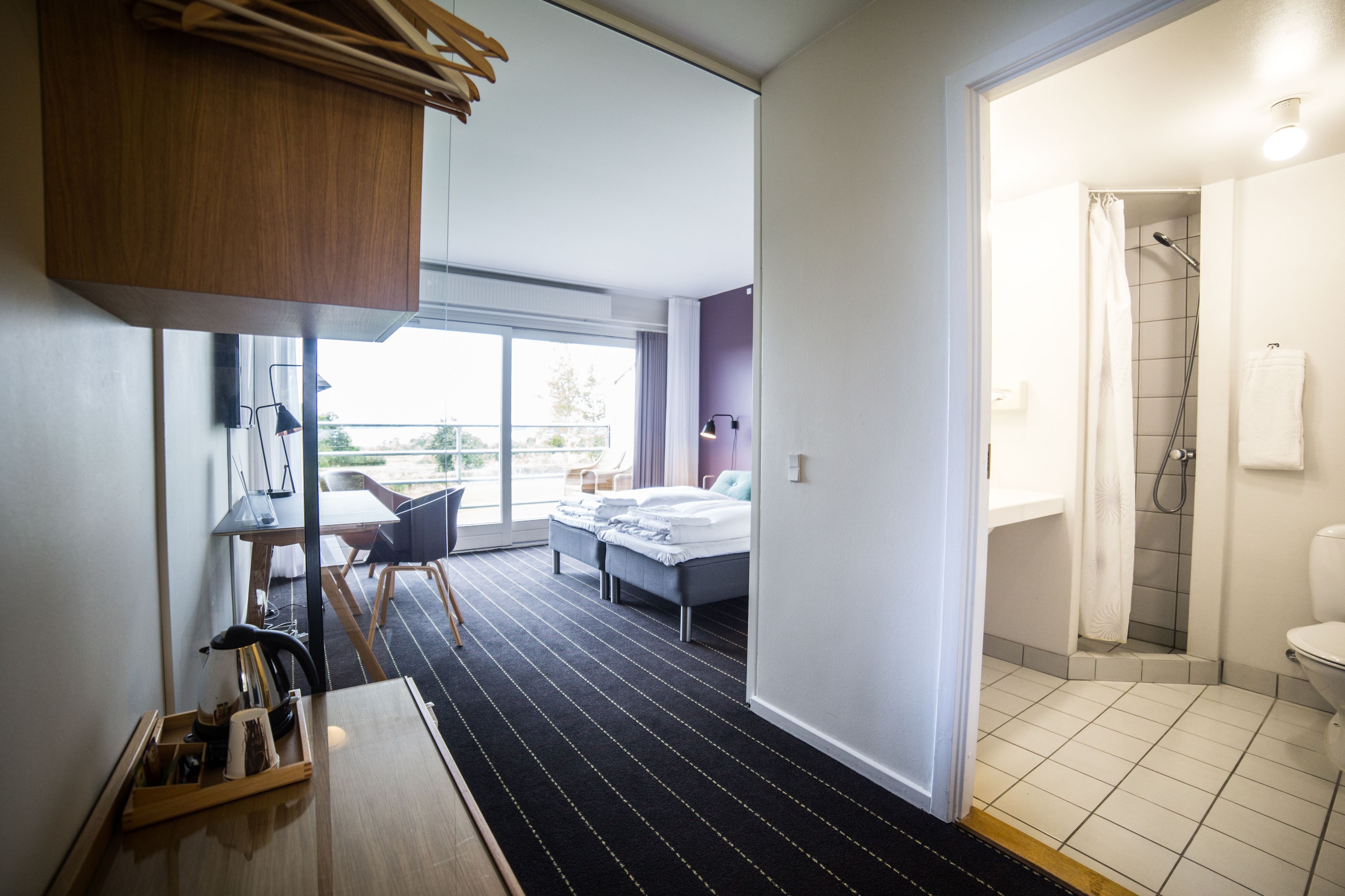 A modern hotel room with a large window, a bed, a desk with a kettle, and a shower area.