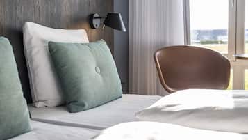 Habitación de hotel con una cama, cabecera de madera, almohada verde y blanca, una silla marrón y una ventana con cortinas.
