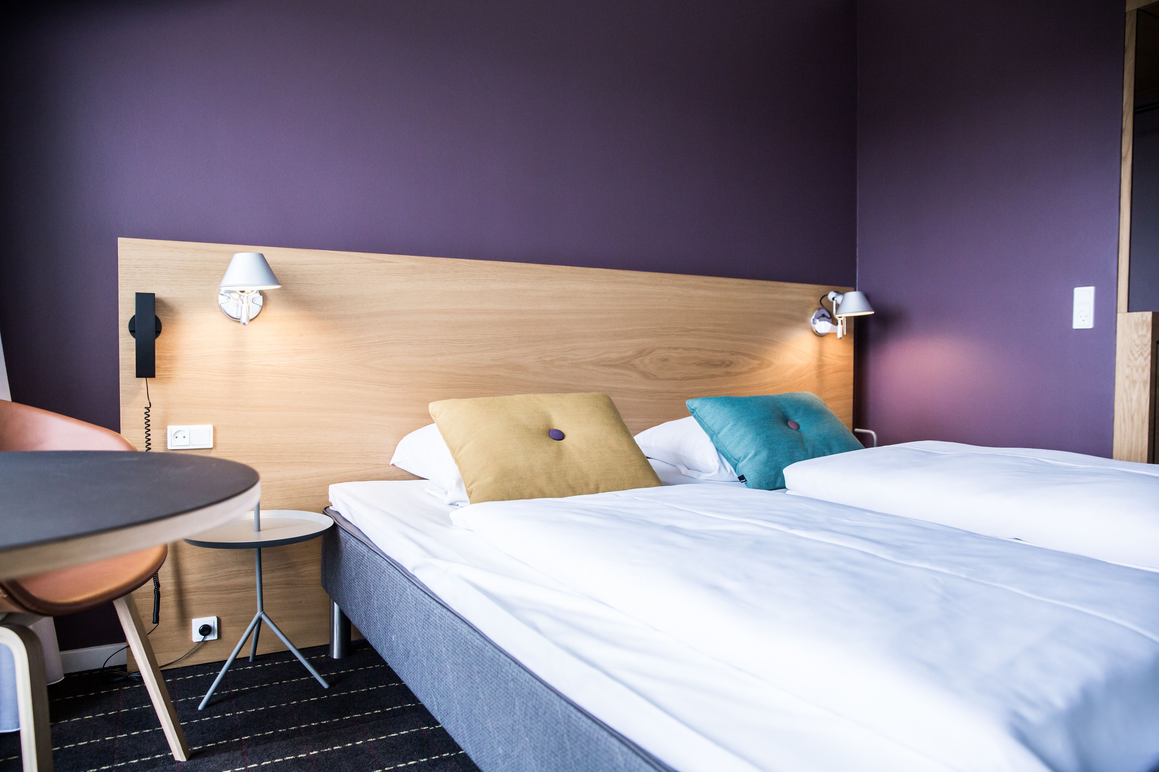 A hotel room with a wooden headboard, a bed with white bedding, a small round table, and a chair.