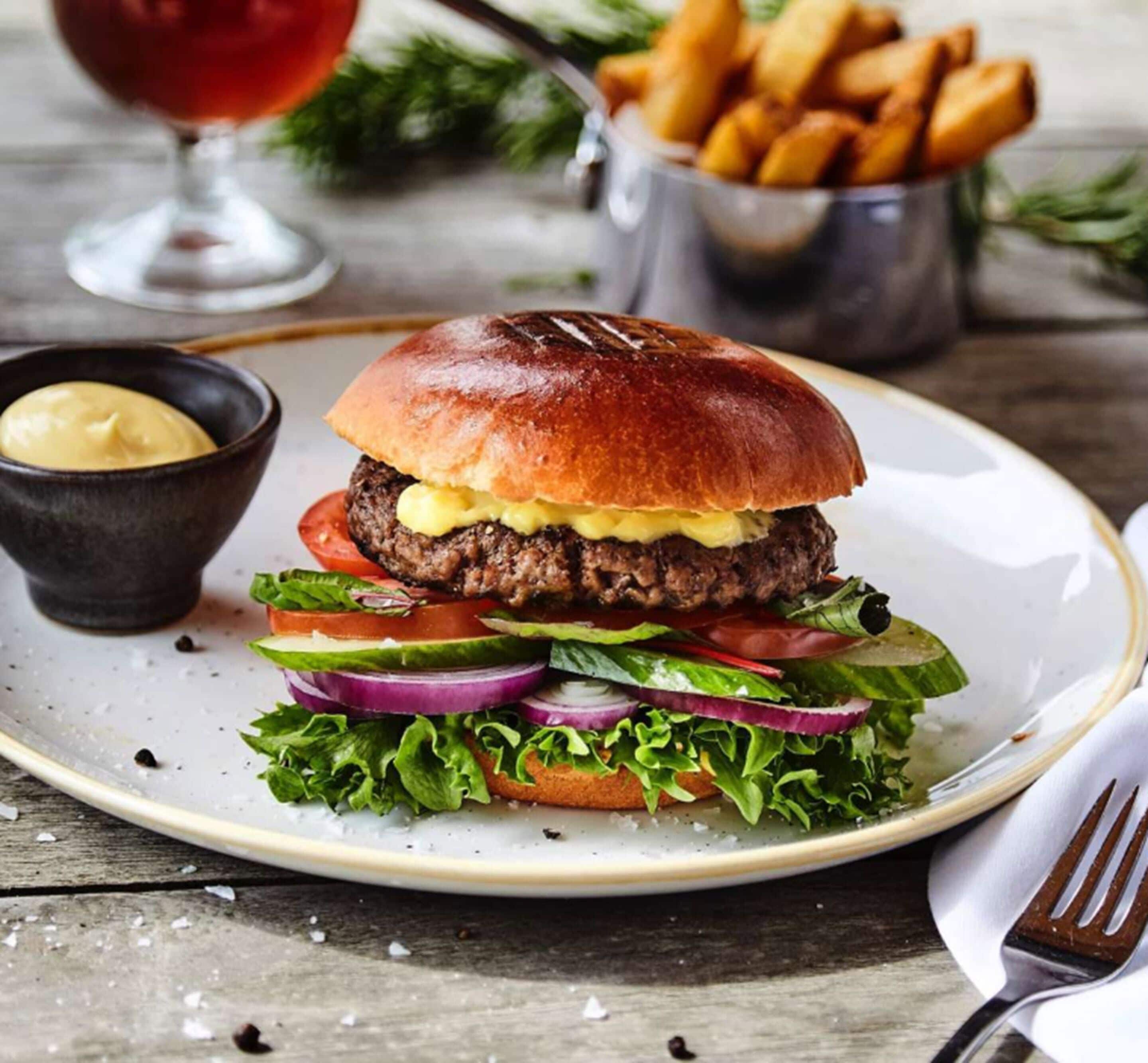 A burger with lettuce, tomato, and red onion on a plate, accompanied by fries and a small bowl of sauce.