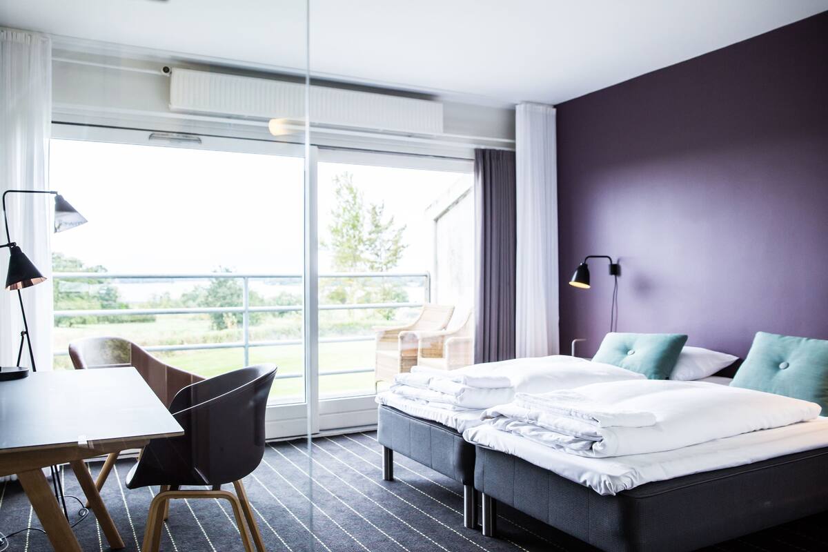 A modern hotel room with a large window, a bed with white linens, a desk with a lamp, and a chair.