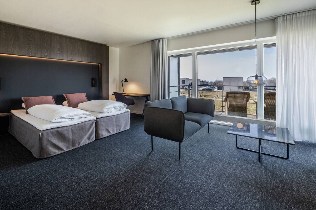 A modern hotel room with a large bed, a sofa, a small table, and a view of the city through large windows.