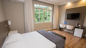 A hotel room with a bed, a desk, a chair, a television, and a window with a view of greenery. - Hotel Himmelblau (Blumenau)