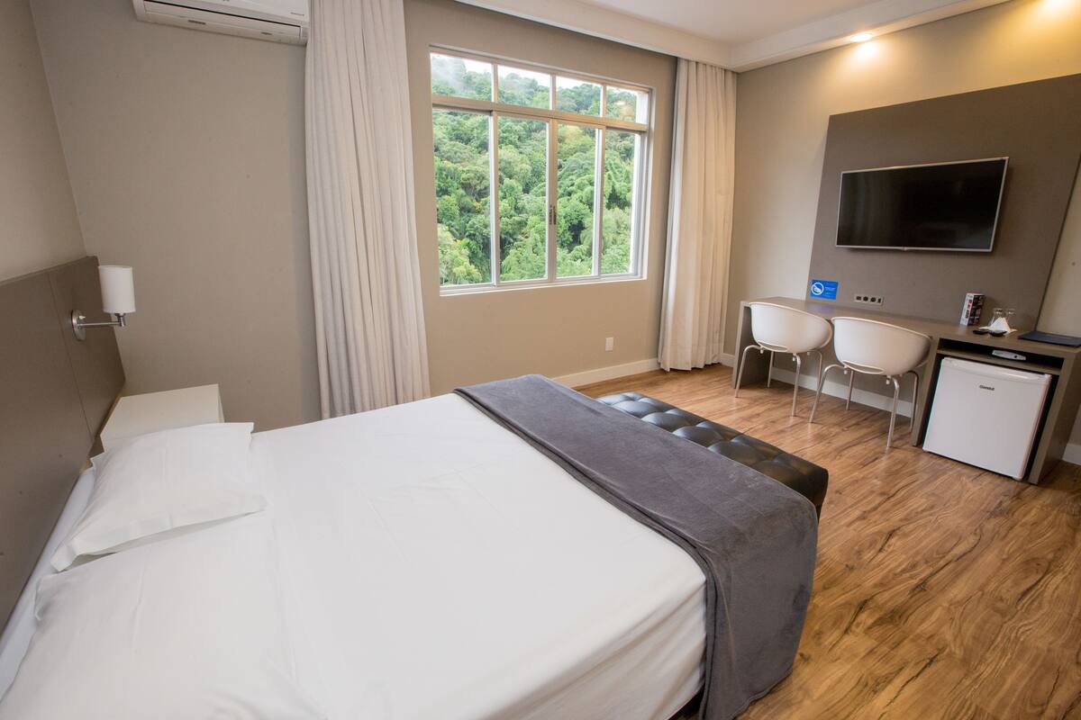 Habitación de hotel con cama, escritorio, silla, televisor y ventana con vista a follaje.