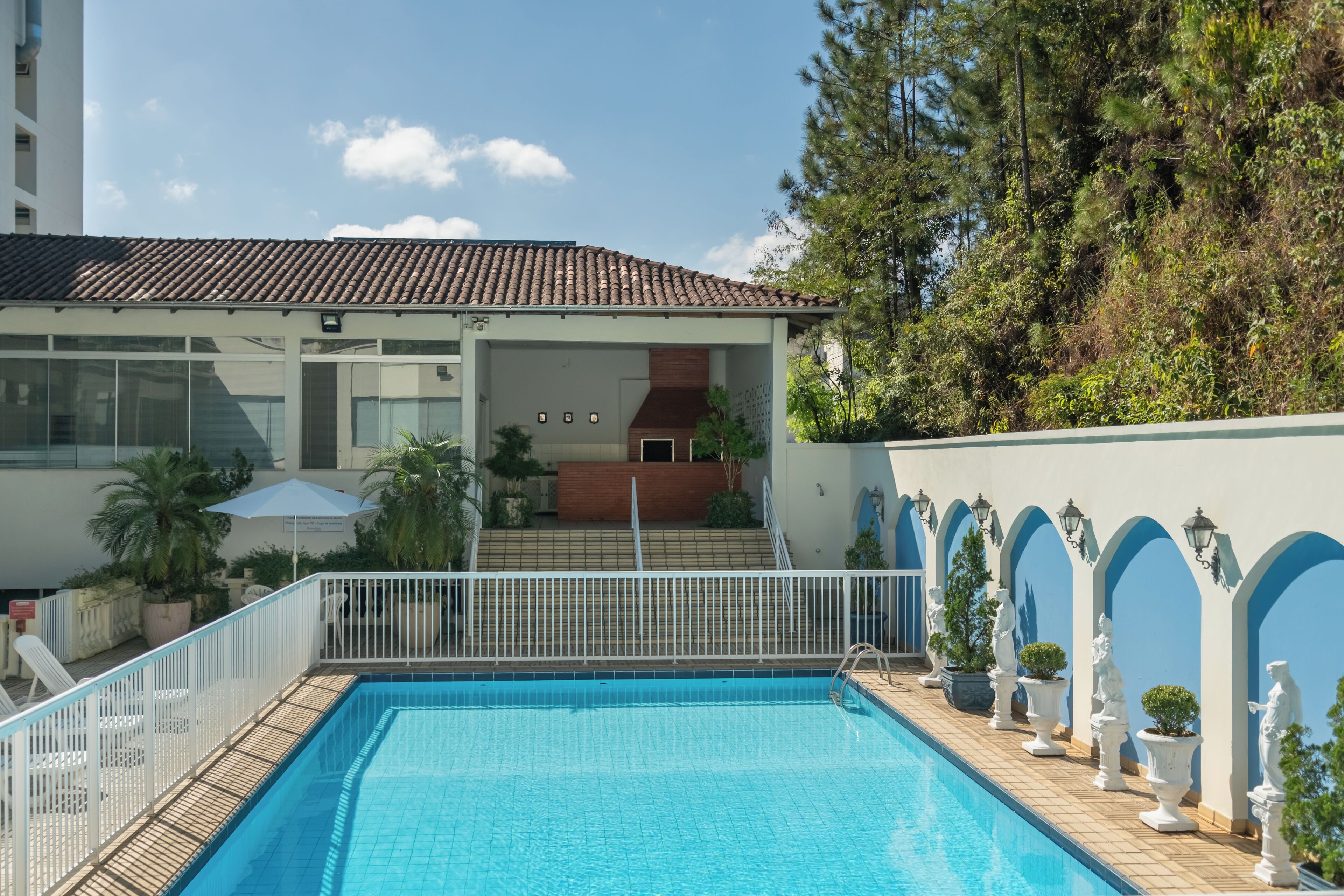 A swimming pool with a tiled deck, surrounded by a white fence and potted plants.