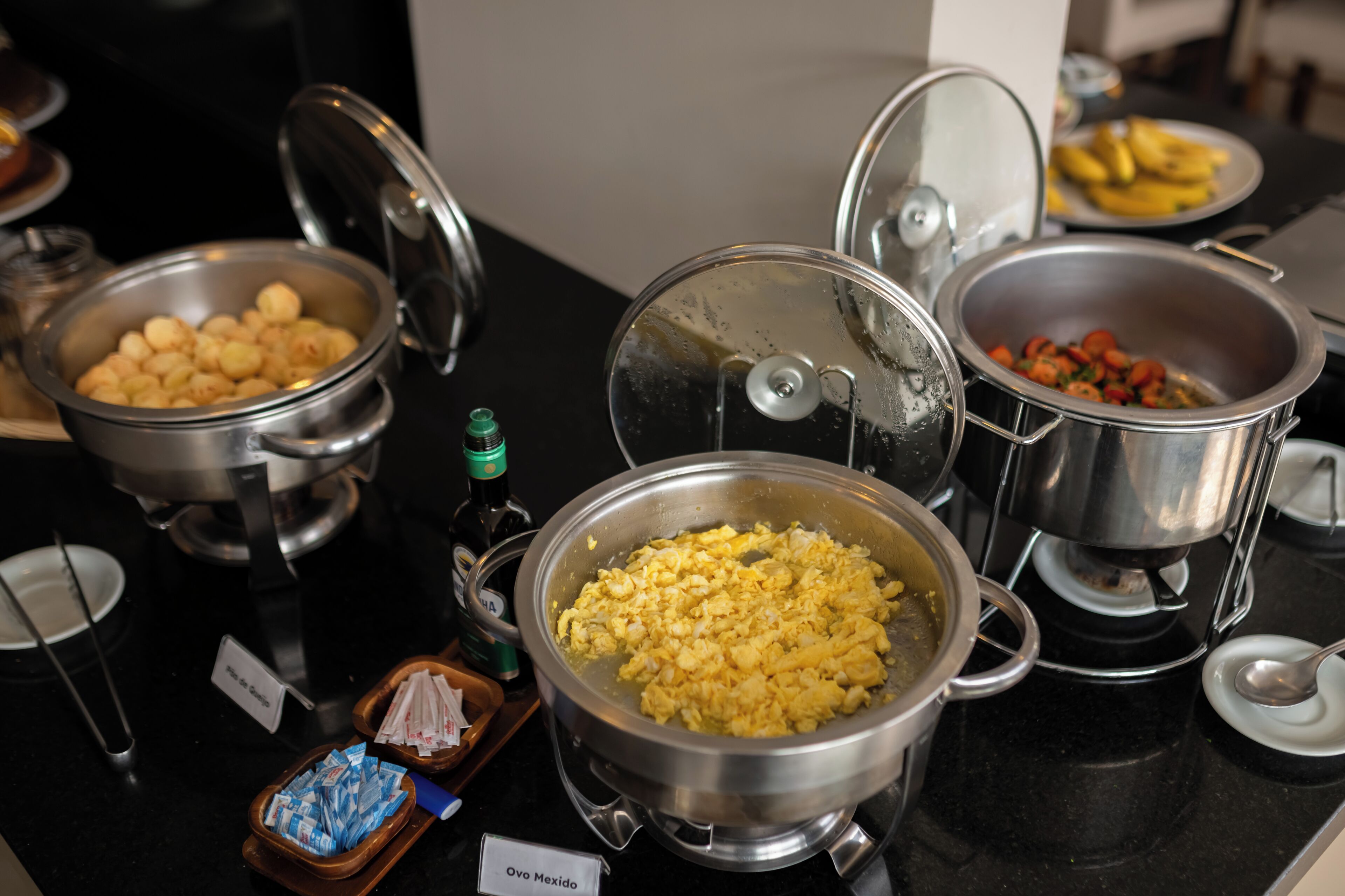 A buffet with various dishes including scrambled eggs, a dish labeled "Ovo Mexido," and a plate of bananas.