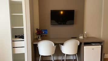 Una habitación compacta con escritorio, dos sillas, un refrigerador pequeño y un televisor montado en la pared.