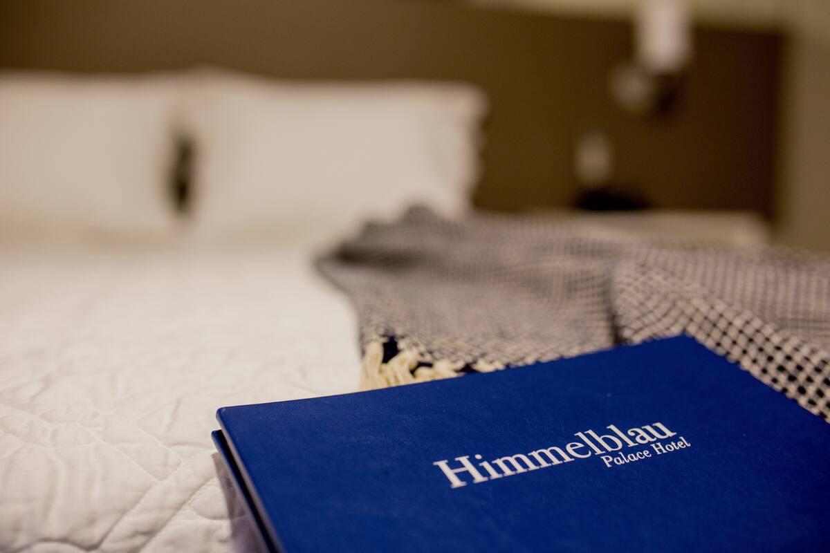 Un libro azul con el título 'Himmelblau Palace Hotel' sobre una cama con colcha blanca.