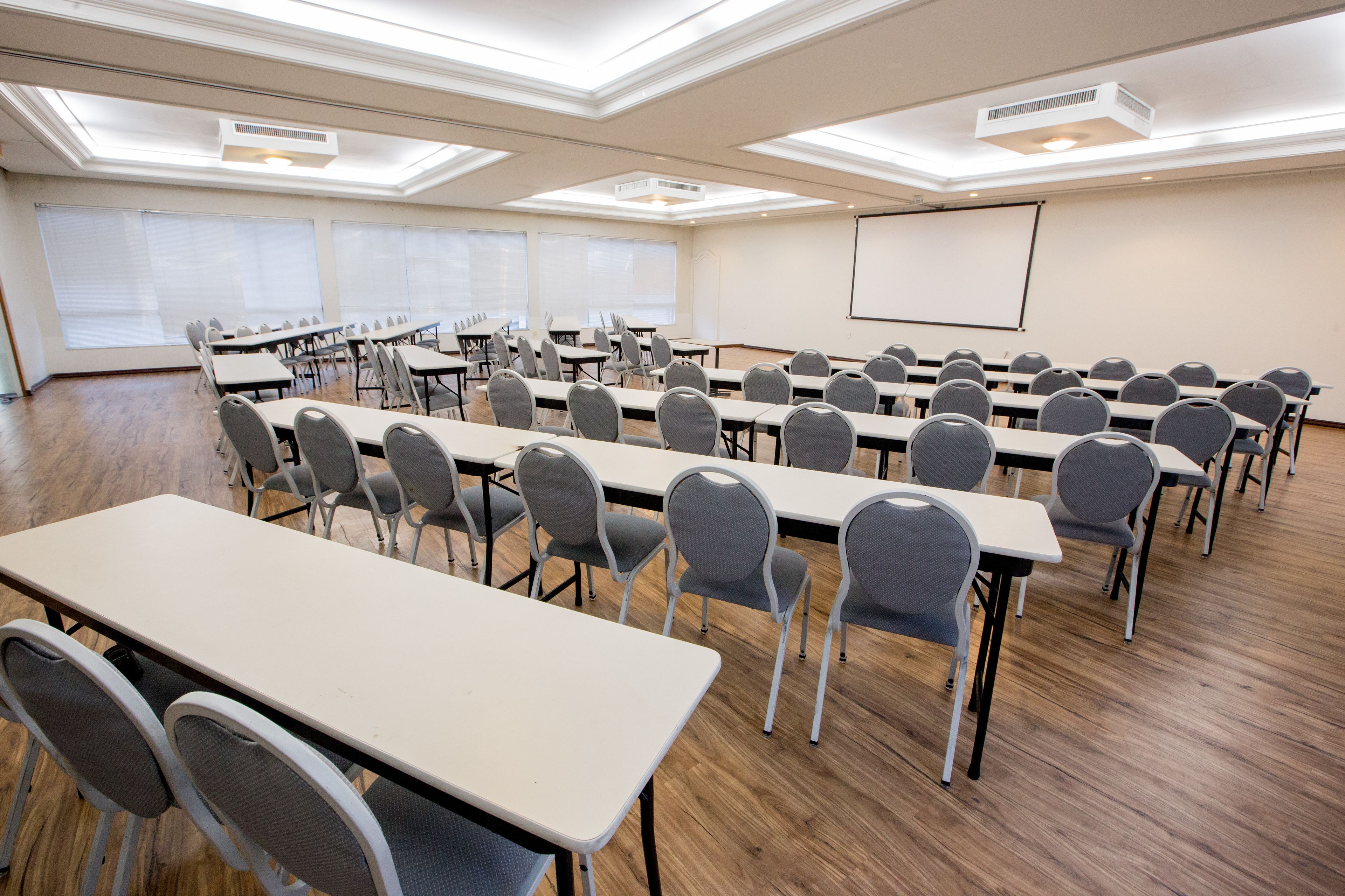 Un salón de conferencias con largas mesas y sillas, una pantalla de proyector y ventanas amplias.