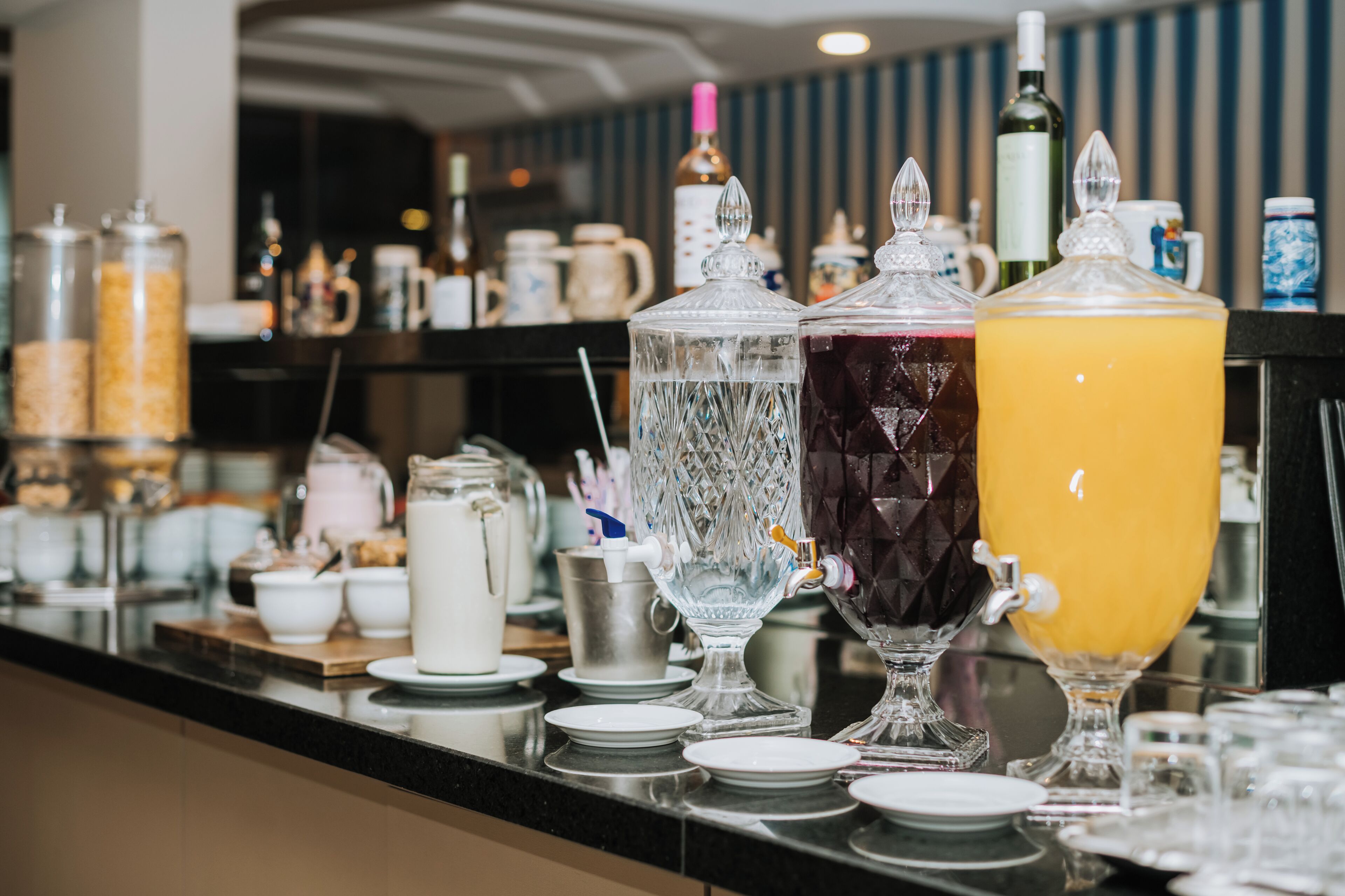 Montaje de bufé con diferentes dispensadores de comida y bebida, incluyendo un dispensador grande de jugo de naranja y varios recipientes de vidrio con diversas bebidas.