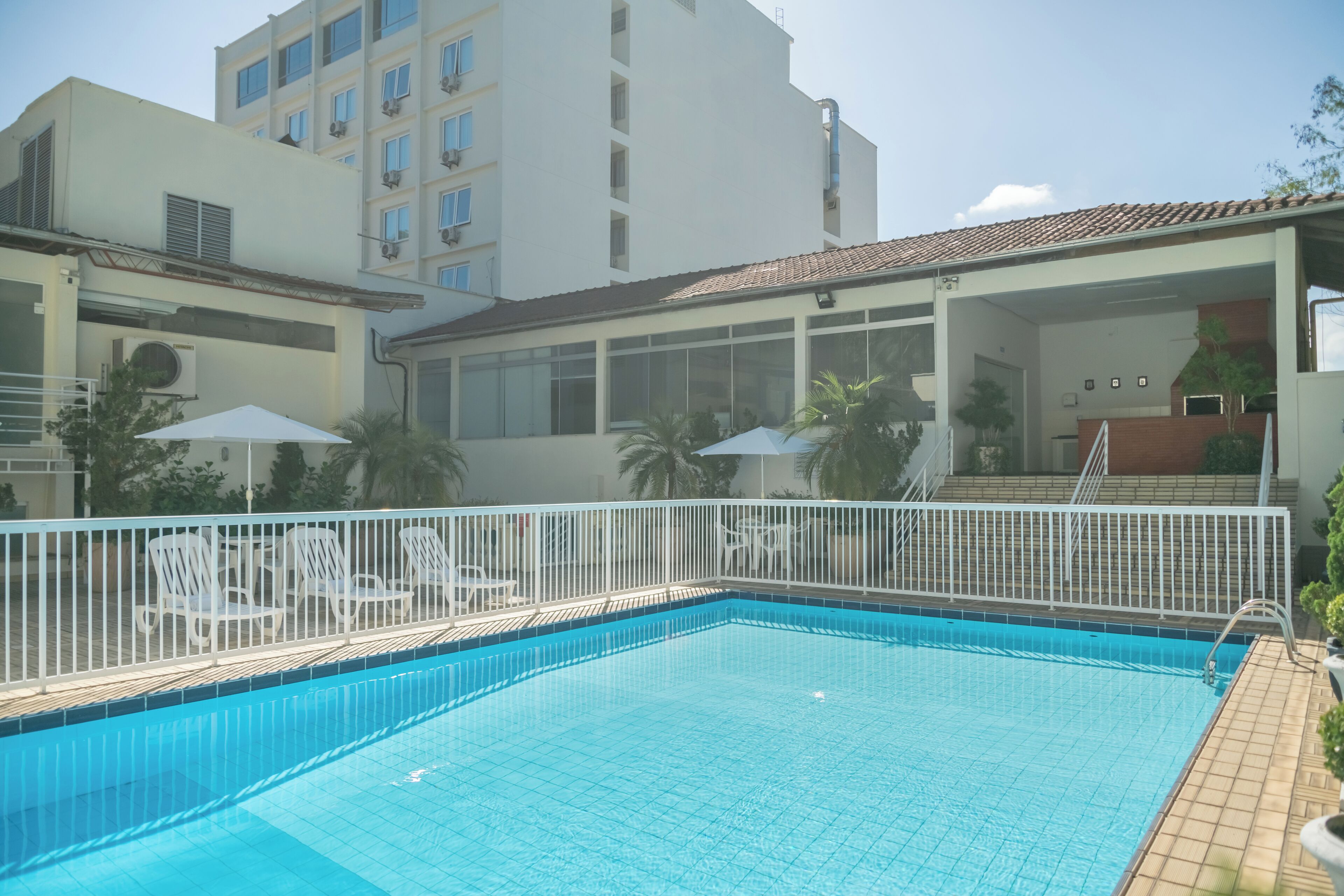Una piscina con bordeado de baldosas, rodeada de sillas de descanso blancas y sombrillas, con un edificio al fondo.
