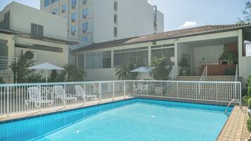 Una piscina con bordeado de baldosas, rodeada de sillas de descanso blancas y sombrillas, con un edificio al fondo.