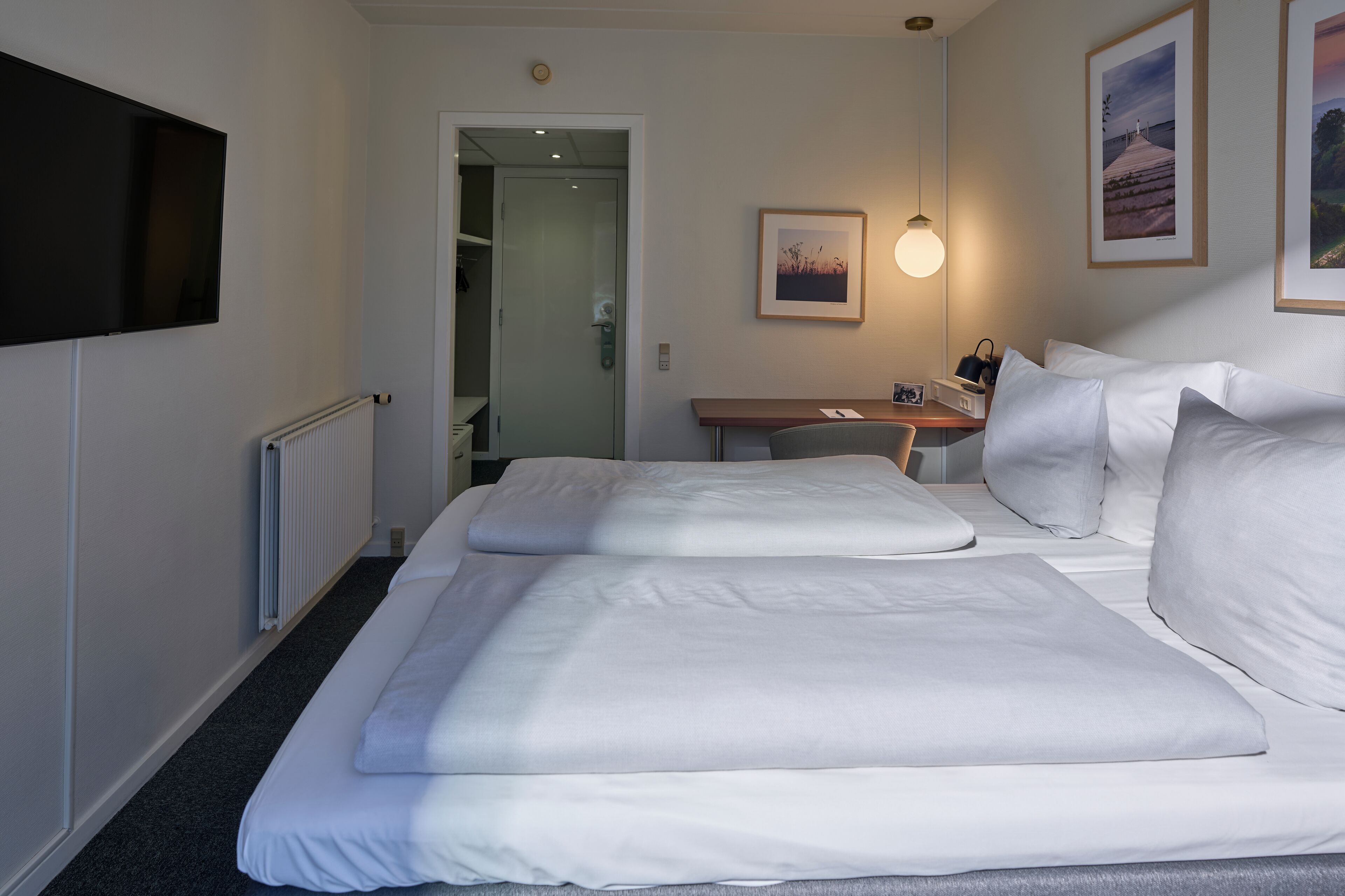 Une chambre d’hôtel avec deux lits, un bureau, une chaise, une télévision et des tableaux encadrés au mur.