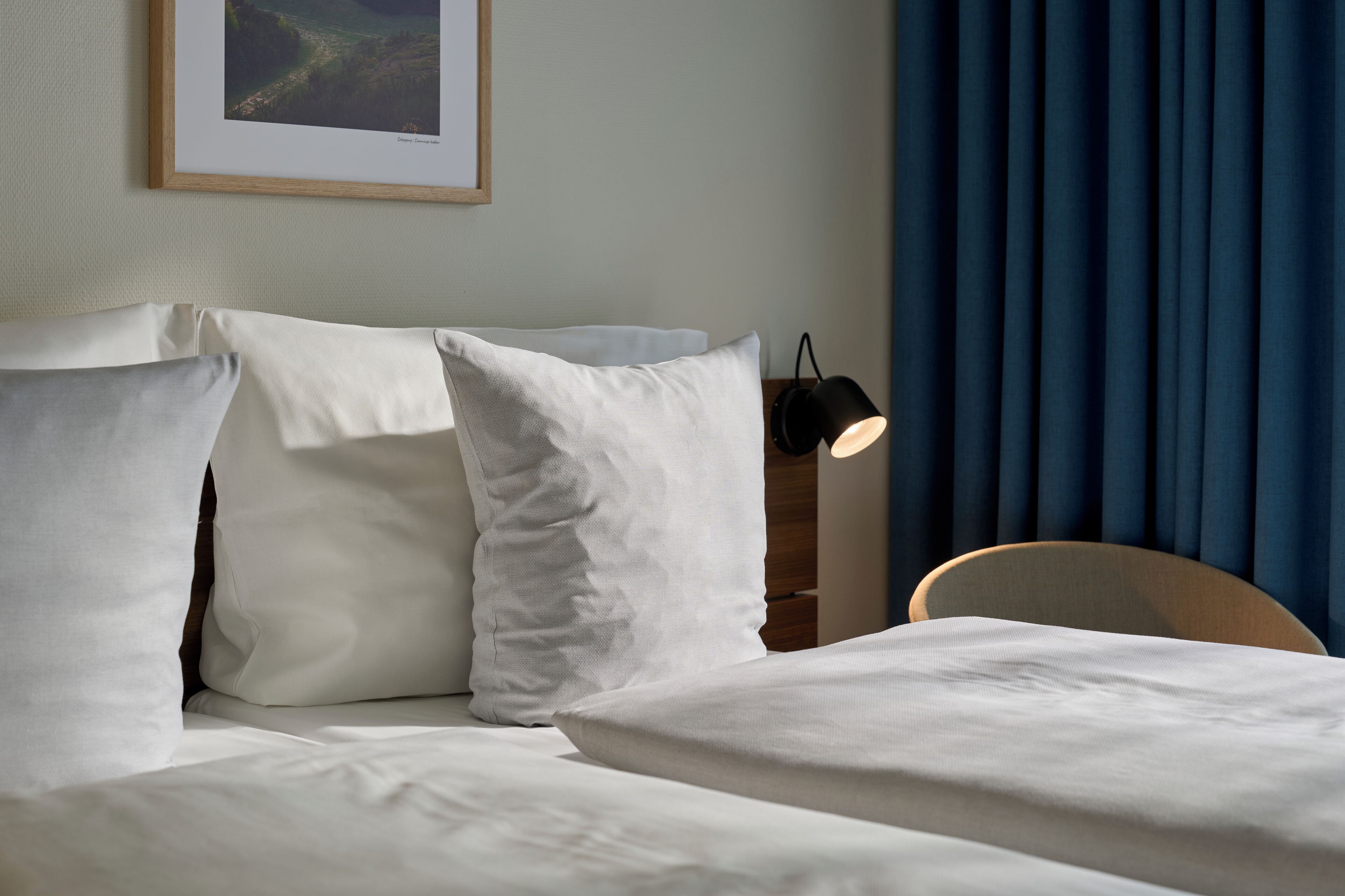 Una cama bien hecha con sábanas blancas, un cuadro en la pared y una lámpara en una mesita de noche.