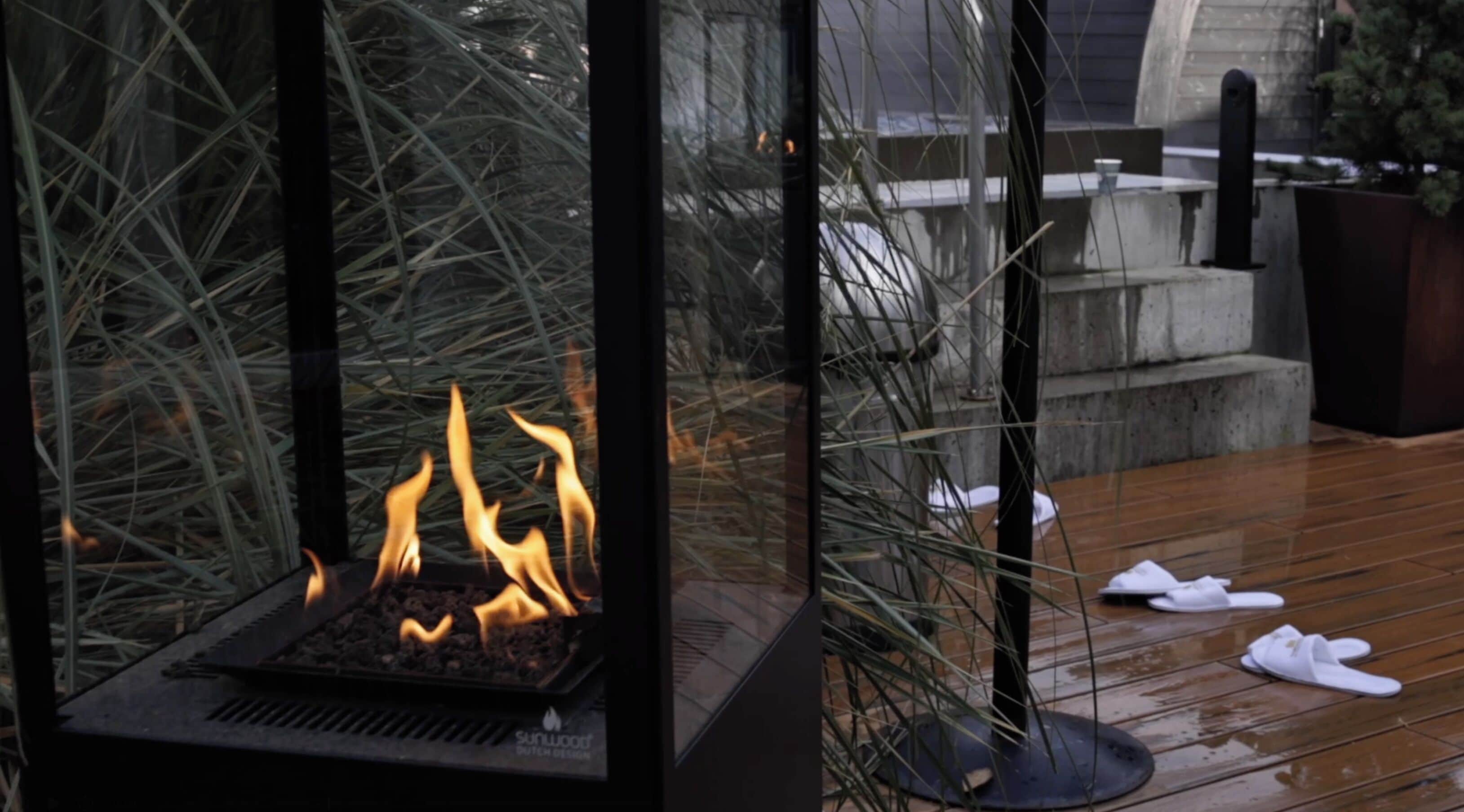 Una chimenea con cristales protectores y llamas visibles, rodeada de pastos altos, sobre una terraza de madera con un banco de piedra y una jardinera.