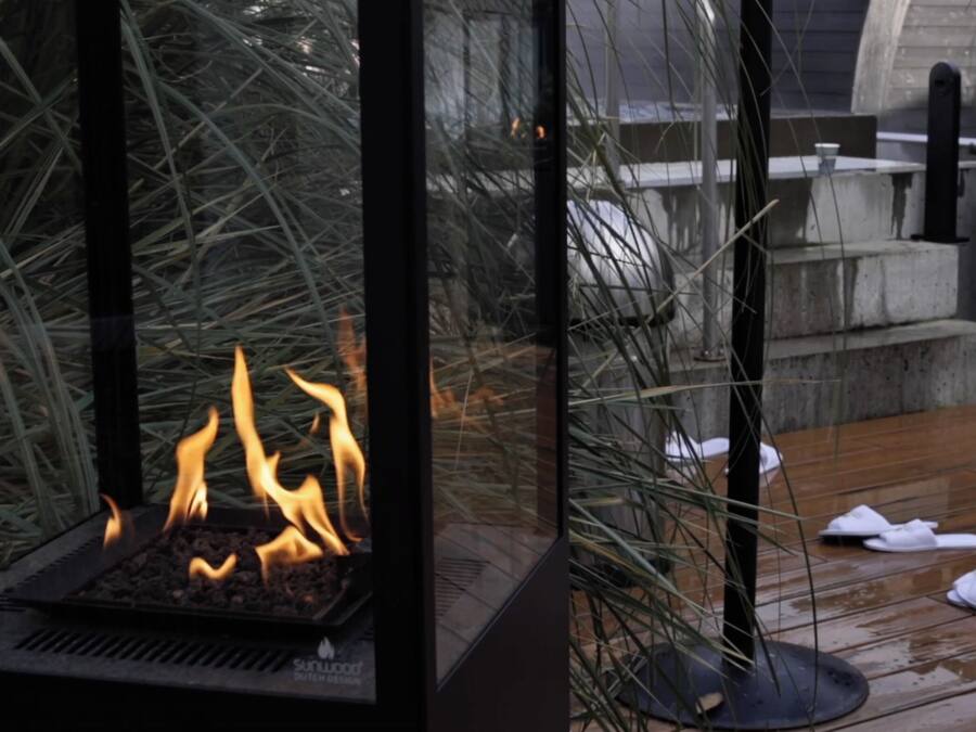 Una chimenea con cristales protectores y llamas visibles, rodeada de pastos altos, sobre una terraza de madera con un banco de piedra y una jardinera.
