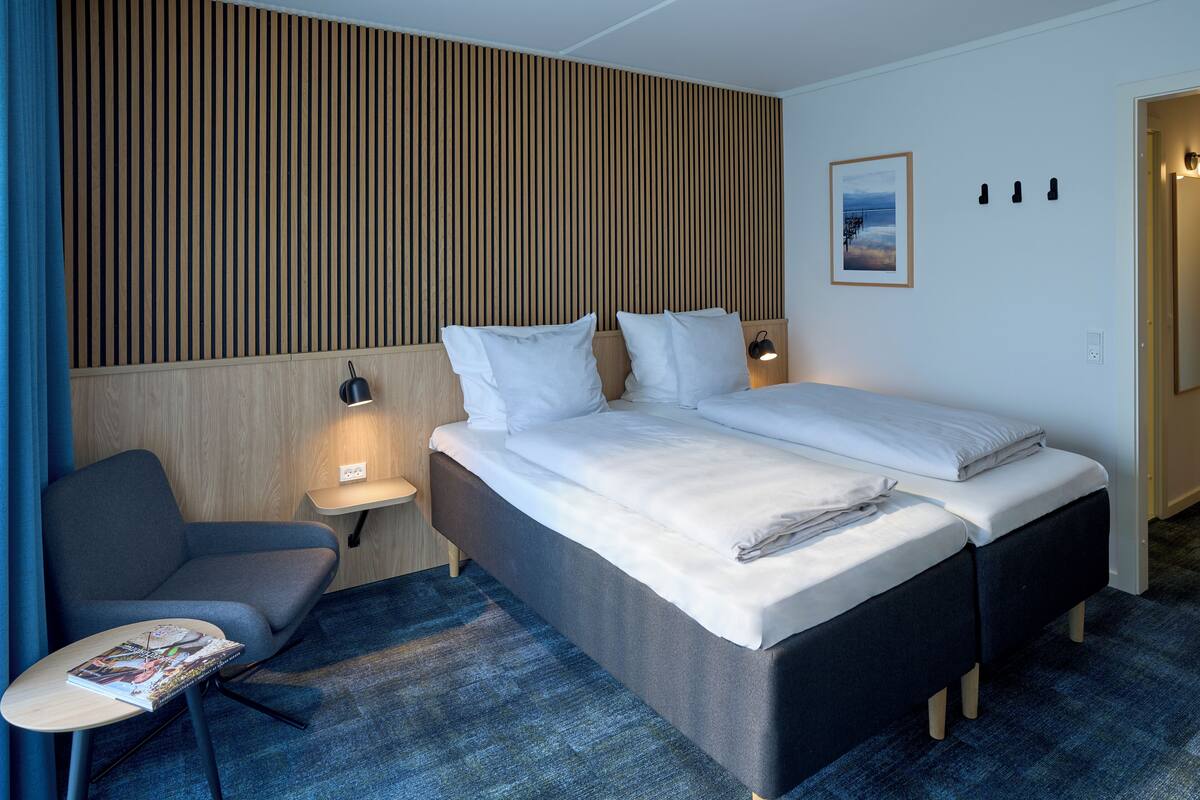 A modern hotel room with a large bed, a bedside table with a lamp, a small desk with a chair, and a view of the sea through the window.