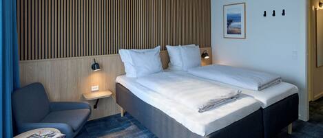 Ein modernes Hotelzimmer mit einem großen Bett, einem Nachttisch mit Lampe, einem kleinen Schreibtisch mit Stuhl und Blick auf das Meer durch das Fenster.