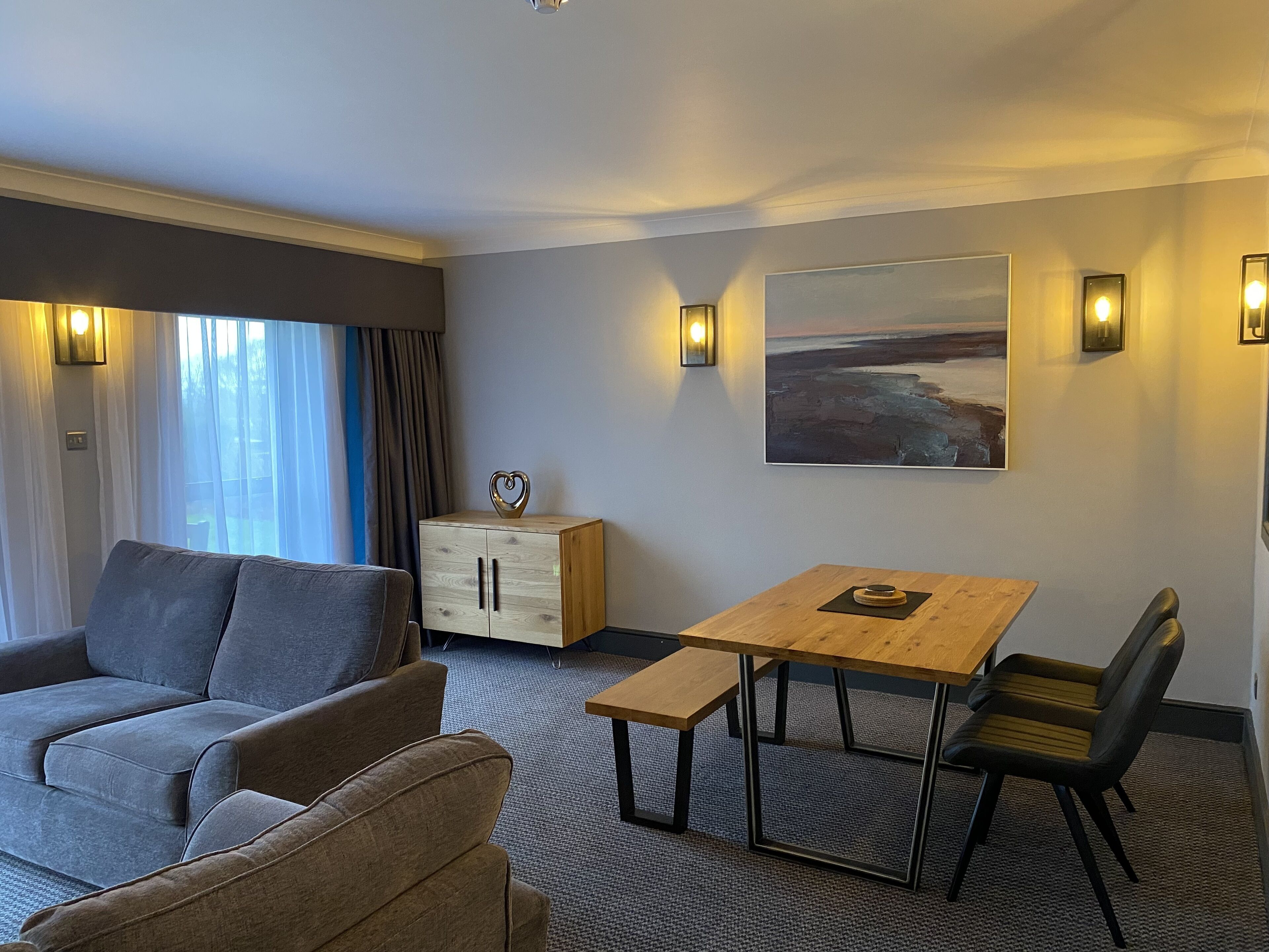 Une chambre d’hôtel moderne avec un canapé gris, une table en bois avec des chaises, un meuble orné d’un motif en forme de cœur et un tableau accroché au mur.