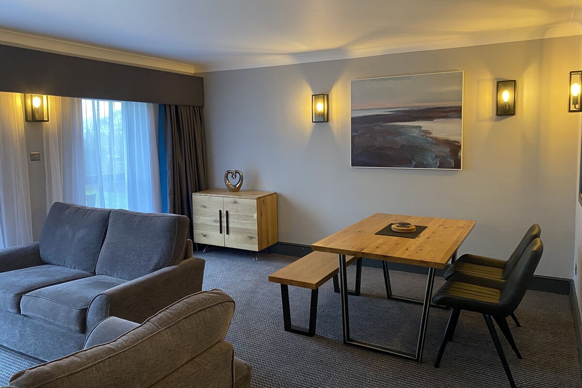 A modern hotel room with a grey sofa, a wooden table with chairs, a cabinet with a heart-shaped decoration, and a painting on the wall.