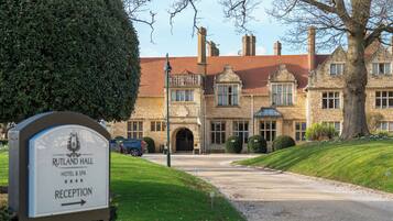 A reception sign for Rutland Hall Hotel & Spa, a stone building with a red-tiled roof, a large tree, and a gravel driveway.