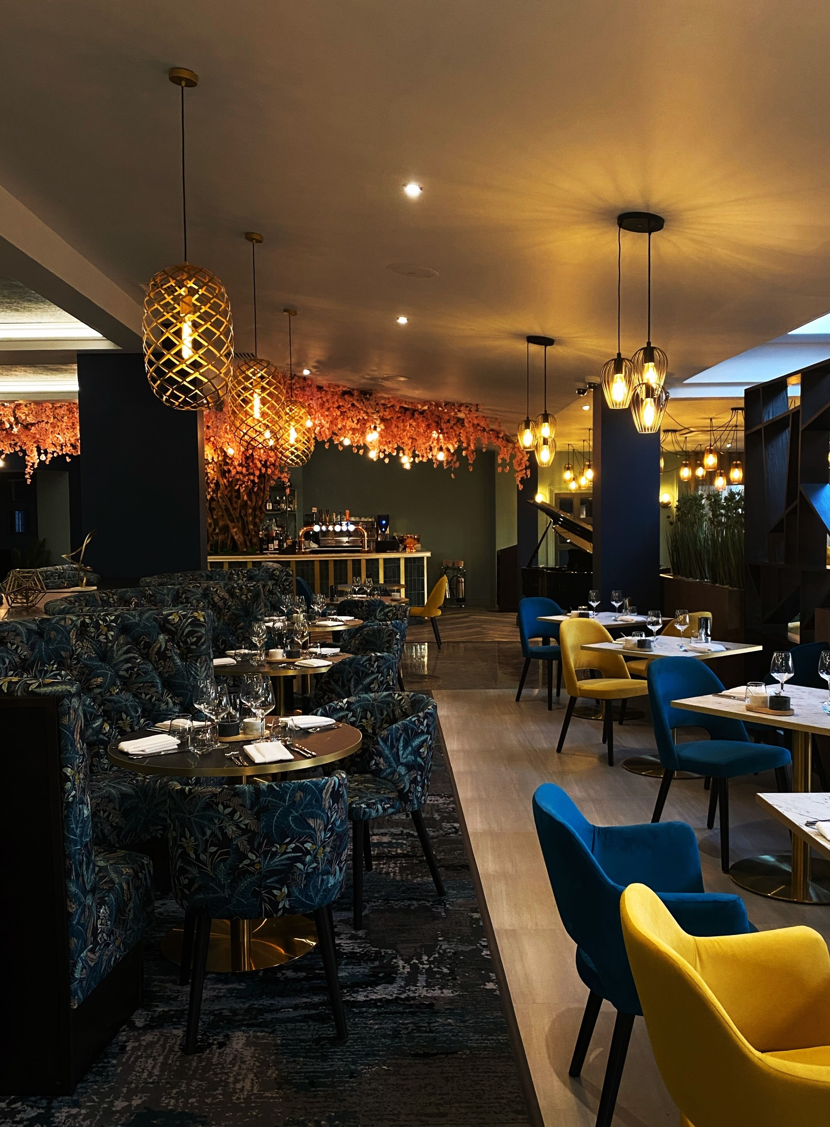 L’intérieur d’un restaurant avec des tables rondes, des chaises bleues et jaunes, des luminaires suspendus et un coin bar.