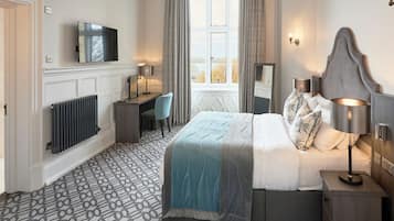 A bedroom with a bed, bedside lamps, a desk, a chair, a radiator, a TV, and a window with curtains.