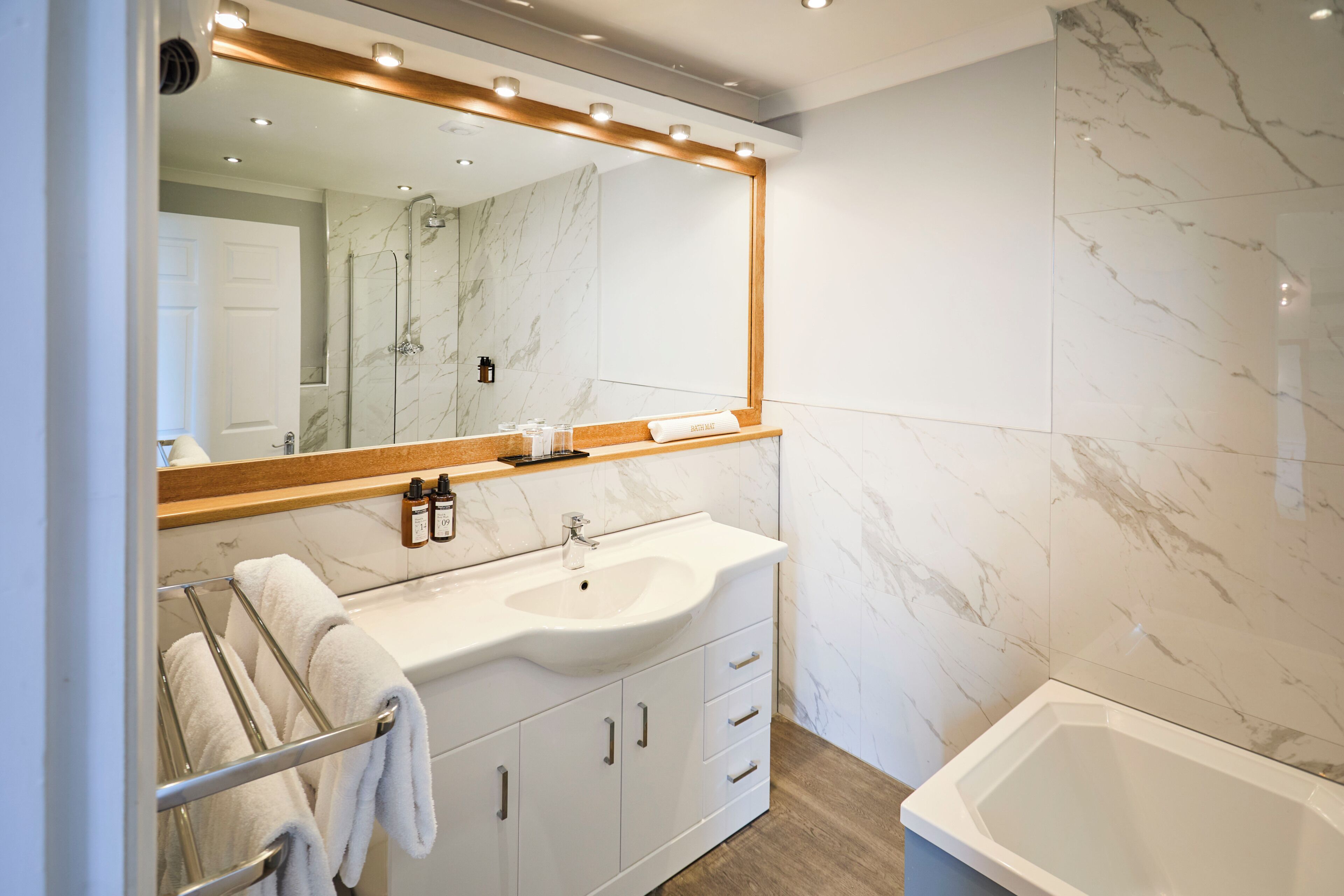 Un baño moderno con lavamanos de mármol, un espejo grande con iluminación y una bañera, además de un perchero para toallas.