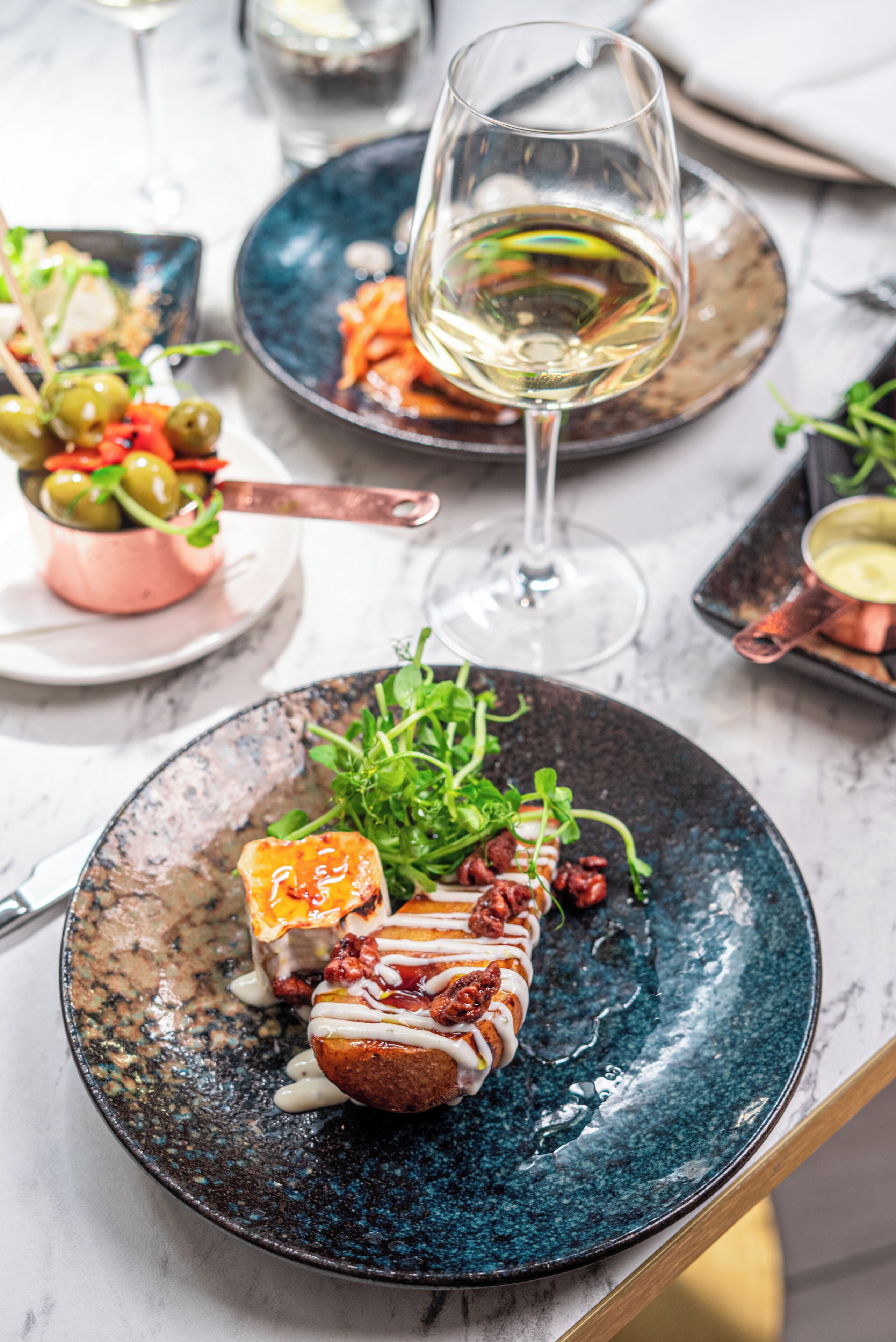 A dining table with various dishes, including a plate with a glazed meat dish, a glass of white wine, and a small bowl of olives.