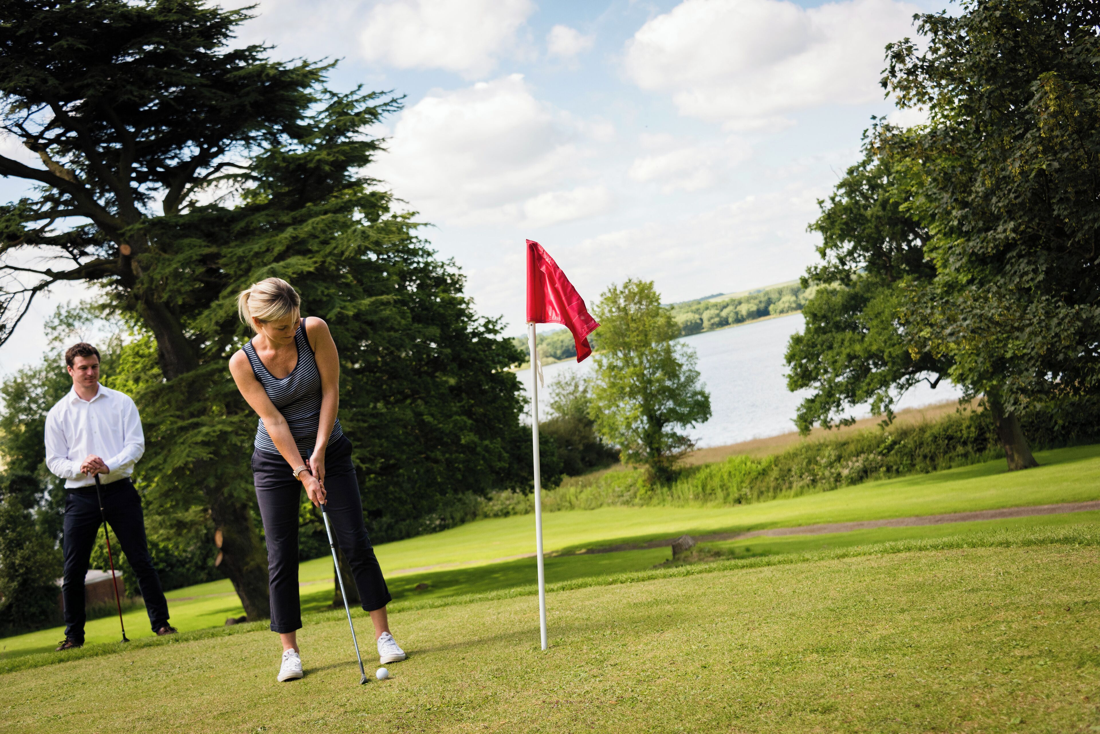 A golf course with a flagstick, a body of water, and trees in the background.