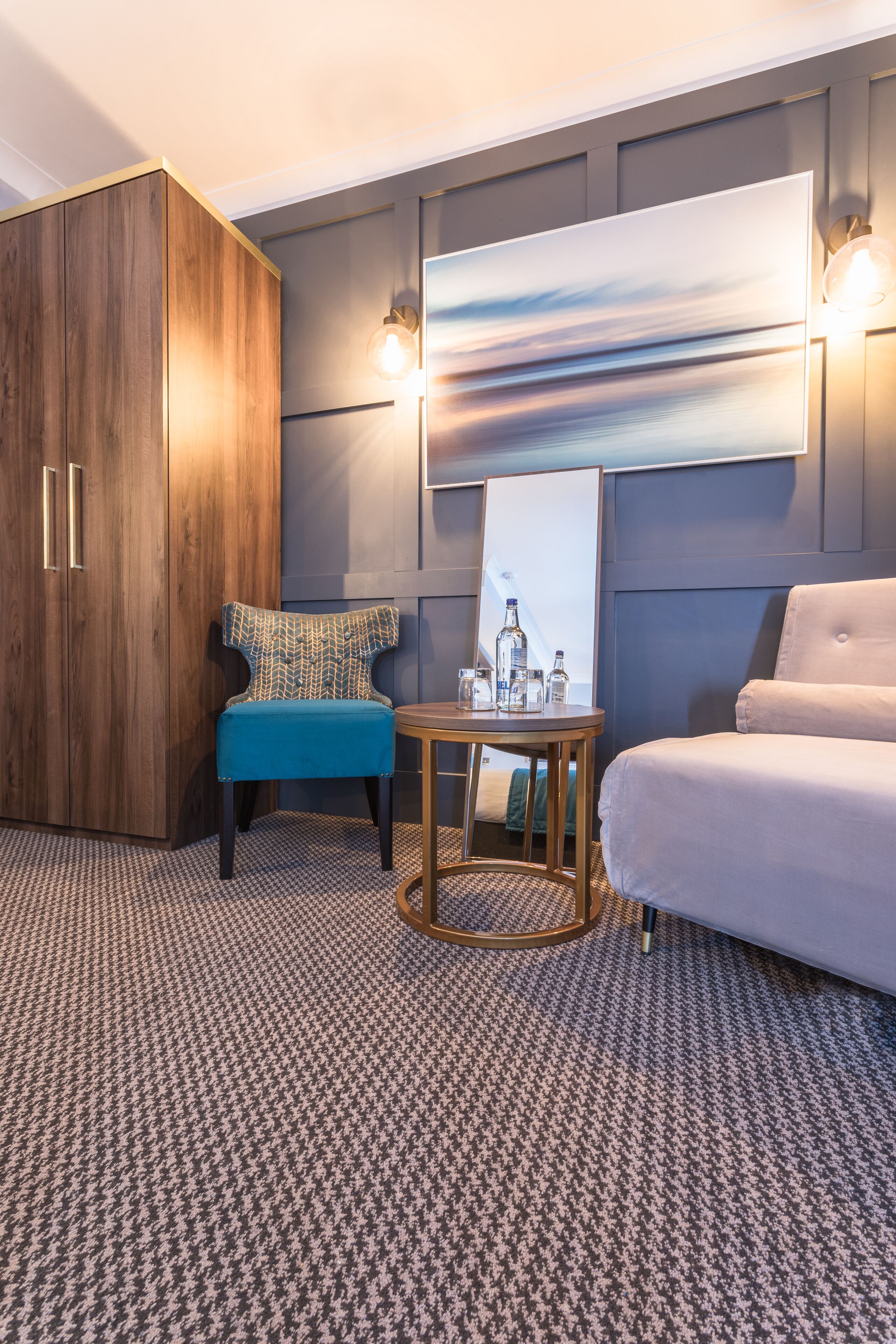 Habitación de hotel con un armario de madera, un sillón azul, una mesa redonda con una botella y un sofá.
