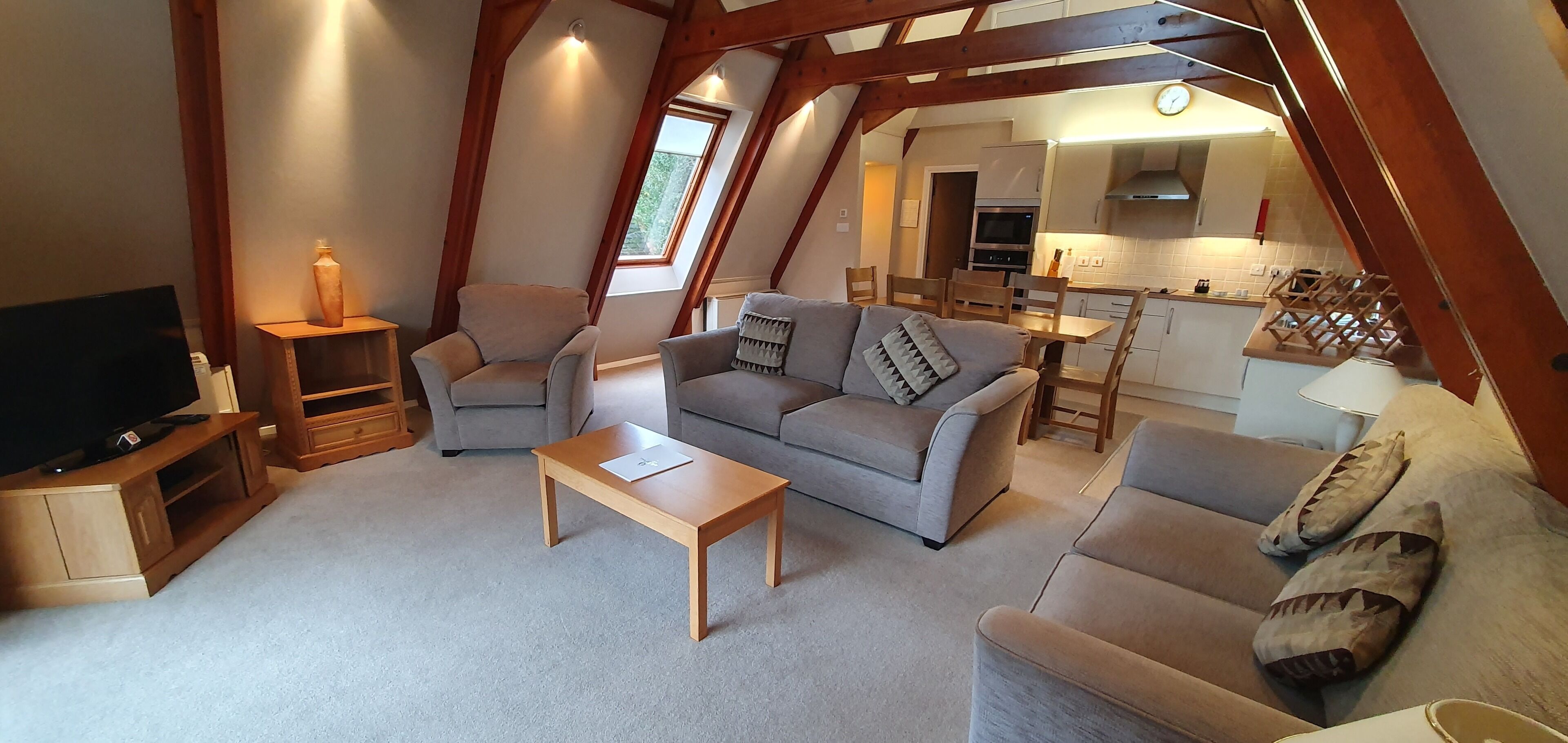 A cozy living room with a sofa, armchairs, a coffee table, and a TV. A kitchen is visible in the background.