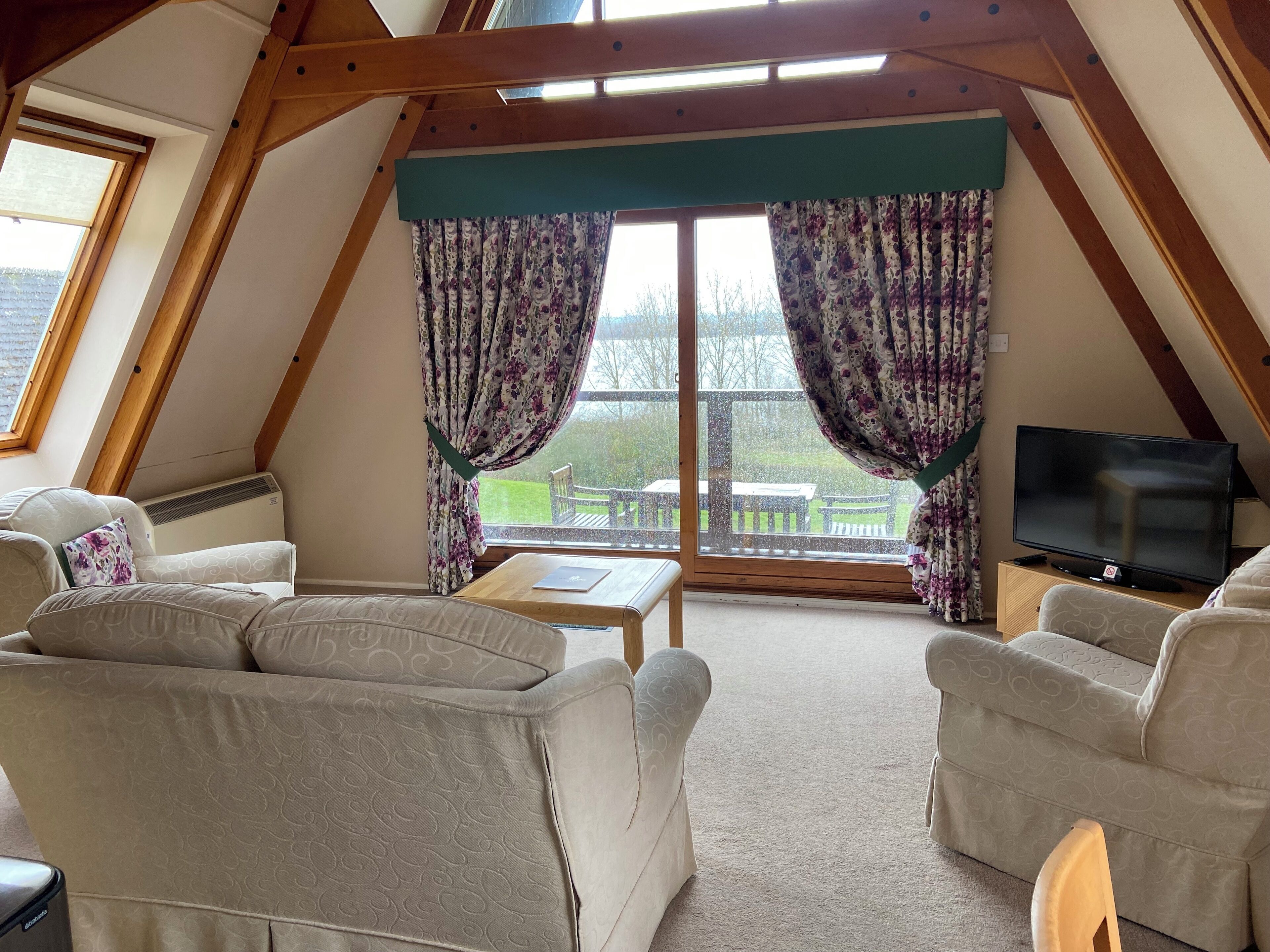 A cozy room with a sloped ceiling, a flat-screen TV, and a view of a green landscape through large windows with floral curtains.