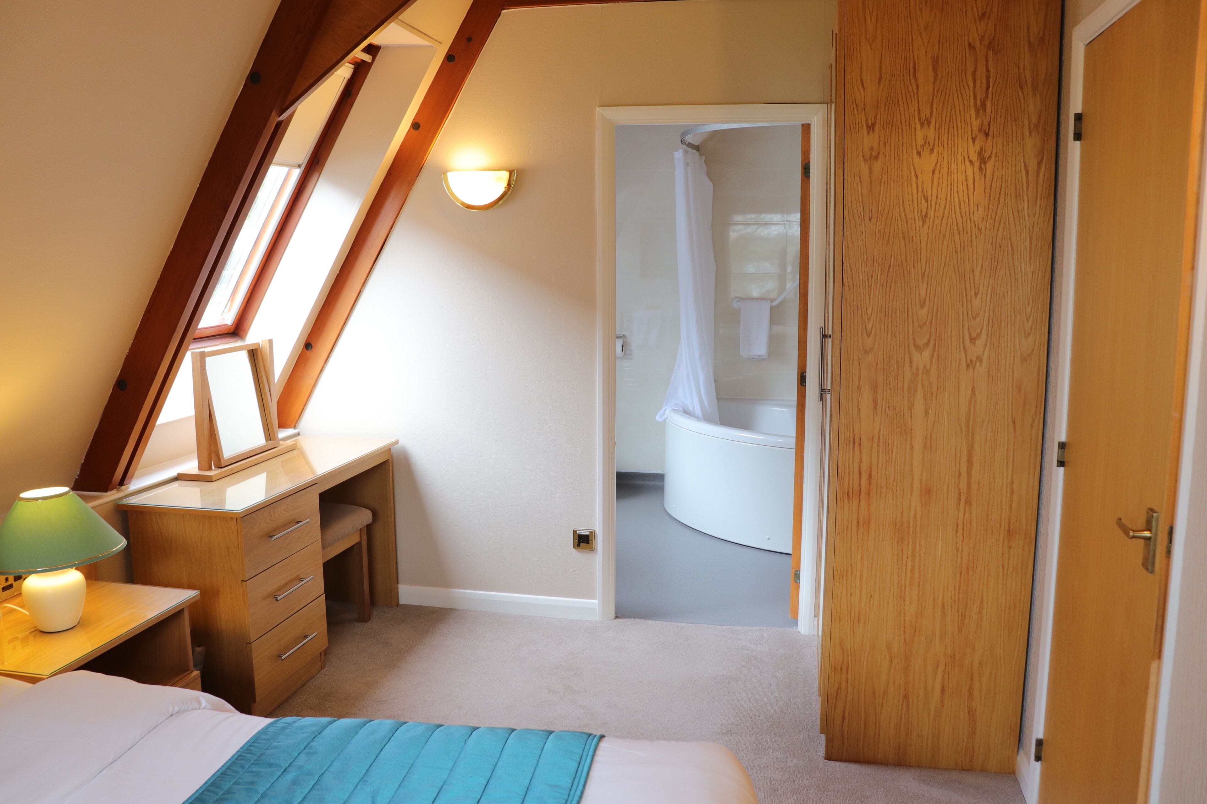 A bedroom with a sloped ceiling, a desk with drawers, a bed with a blue blanket, and a bathroom visible through an open door.