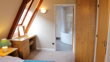 A bedroom with a sloped ceiling, a desk with drawers, a bed with a blue blanket, and a bathroom visible through an open door.