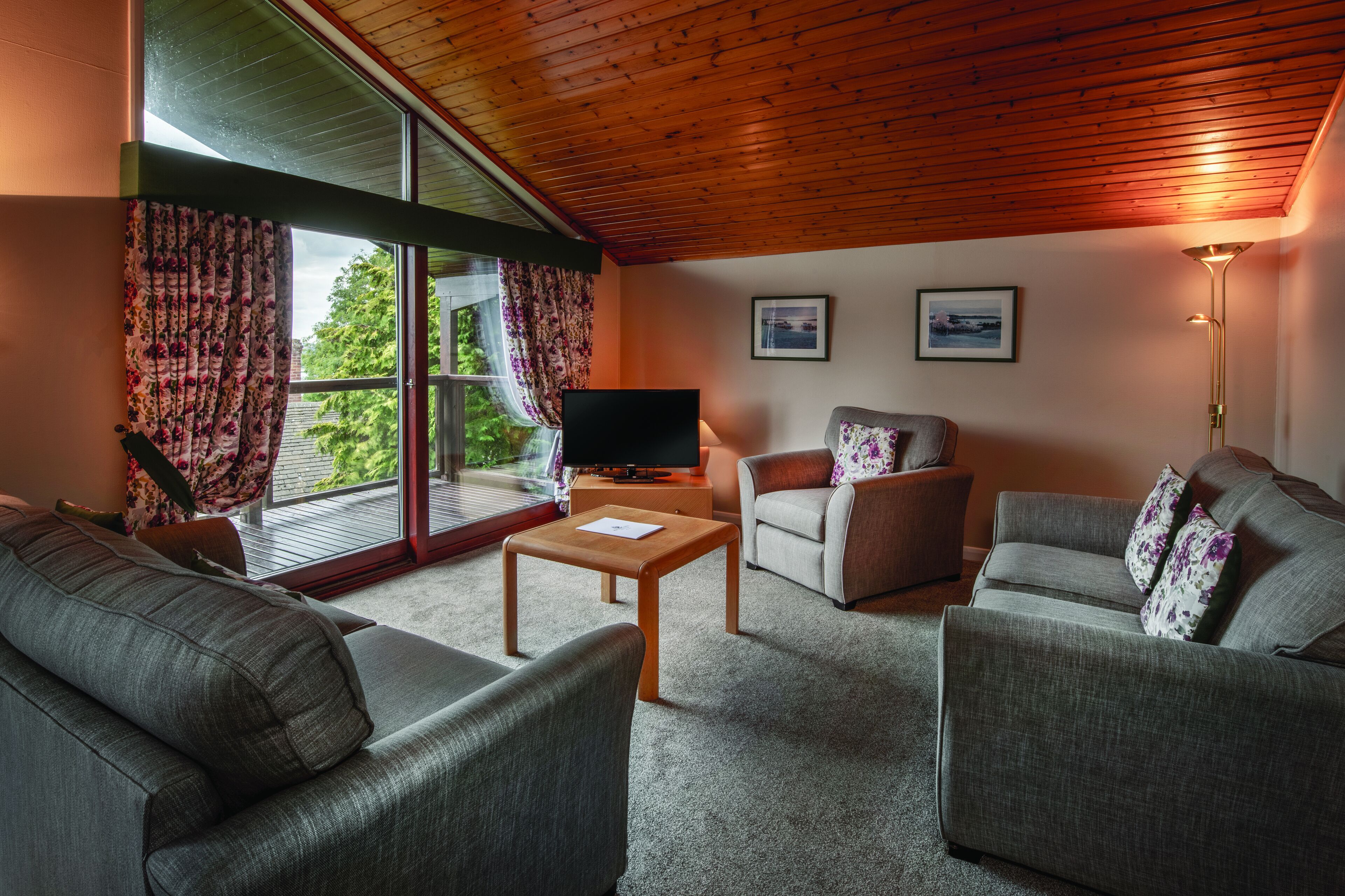 A living room with a wooden ceiling, large windows with curtains, a flat-screen TV, two sofas, a coffee table, and a floor lamp.