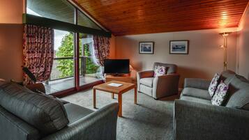 A living room with a wooden ceiling, large windows with curtains, a flat-screen TV, two sofas, a coffee table, and a floor lamp.
