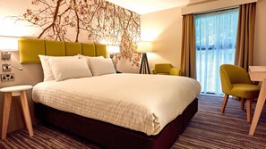 A hotel room with a large bed, a green headboard, two bedside lamps, a small round table, a yellow armchair, and a view of the outdoors through a window with curtains. - Holiday Inn Northampton West M1, Jct 16 by IHG (Northampton)