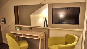Une chambre d’hôtel avec un lit, un bureau, deux chaises jaunes, une télévision et un miroir.