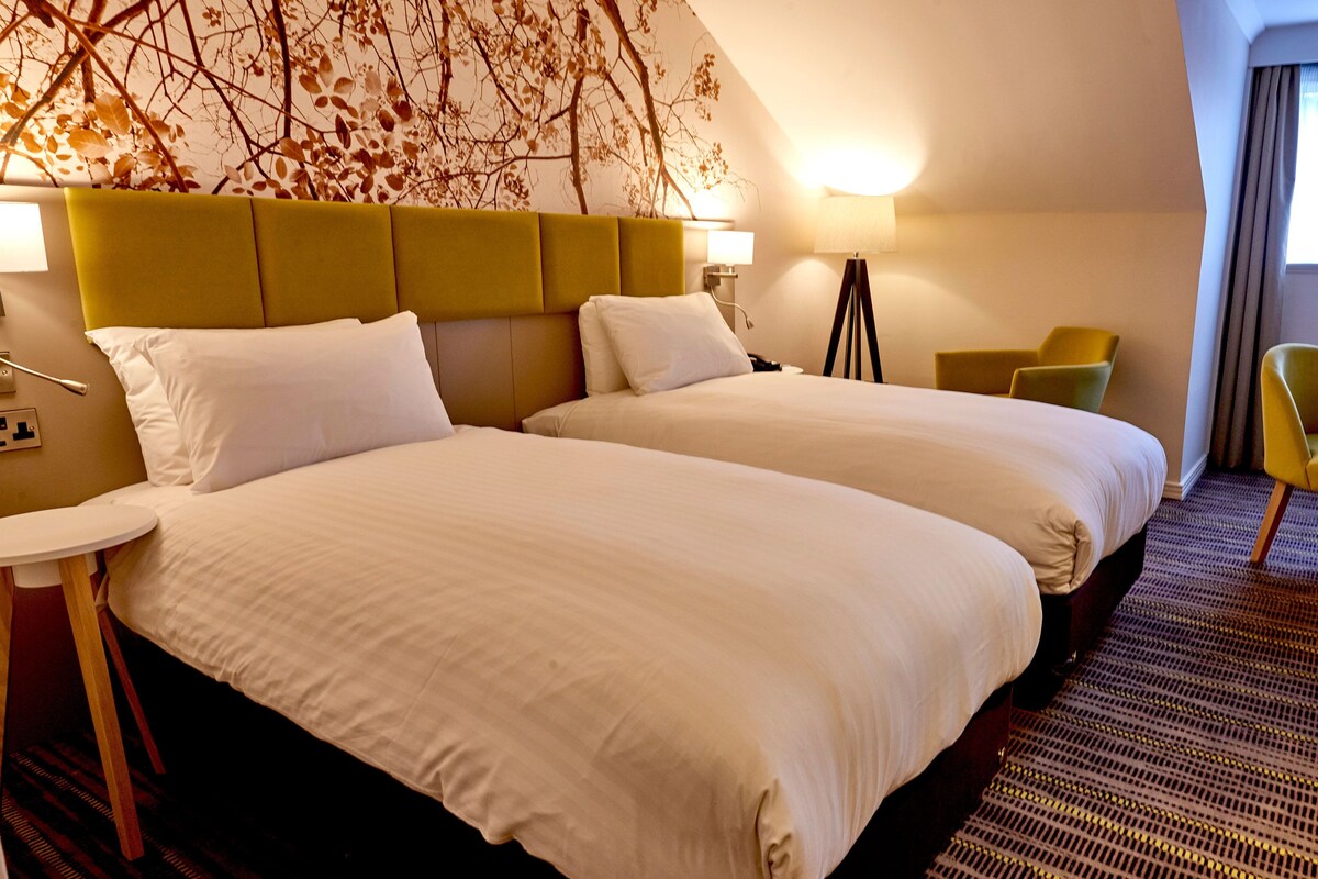 A hotel room with two beds, a desk, a chair, and a lamp.
