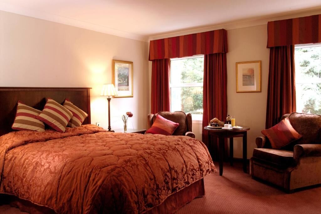 Habitación de hotel con una cama grande, dos sillones, una mesita de noche con lámpara y una ventana con cortinas.
